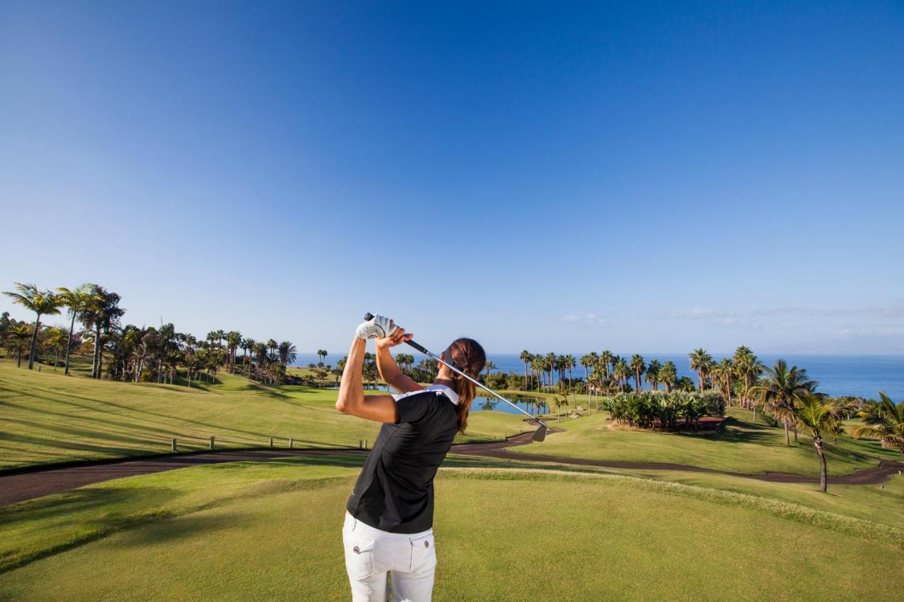 Professional golfer mid-swing on lush fairway with palm trees, Atlantic Ocean and mountains in background