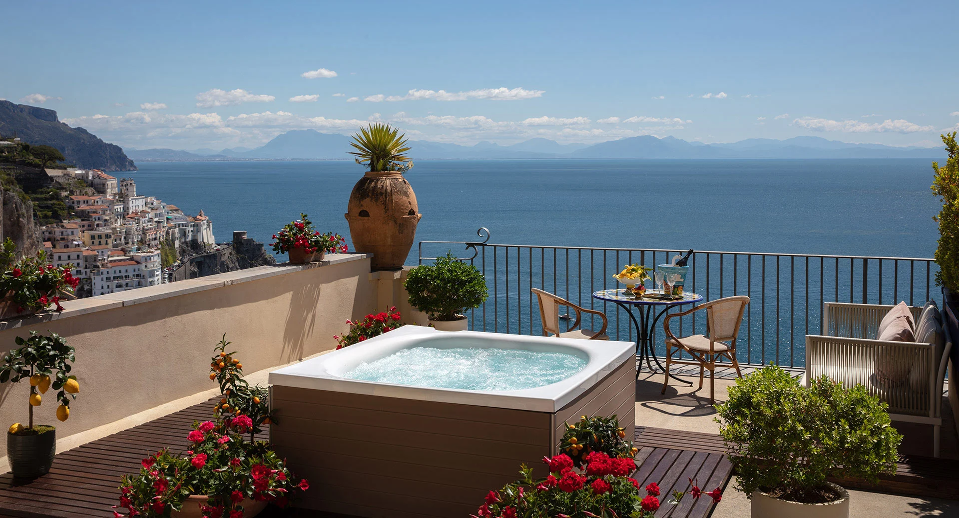 Luxurious cliffside terrace with white soaking tub, decorative terracotta pots, red flowering plants, dining table with sea views, colorful Mediterranean village buildings on cliffs below, and turquoise Amalfi Coast sea