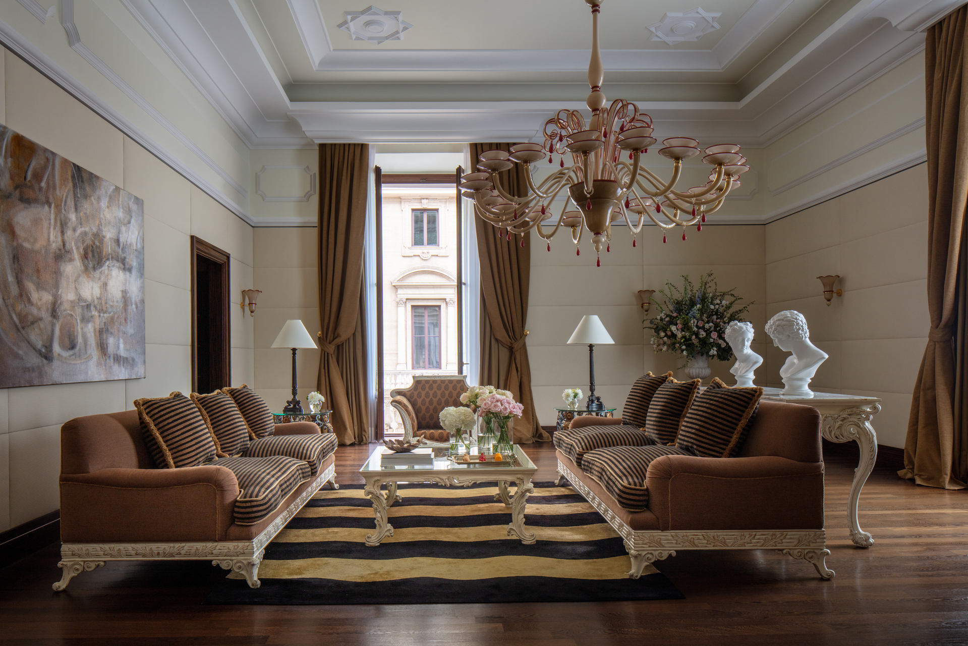 Elegant neoclassical living suite with ornate brown and tan striped sofas, crystal chandelier, tall draped windows, white marble busts, and sophisticated classical Roman luxury design
