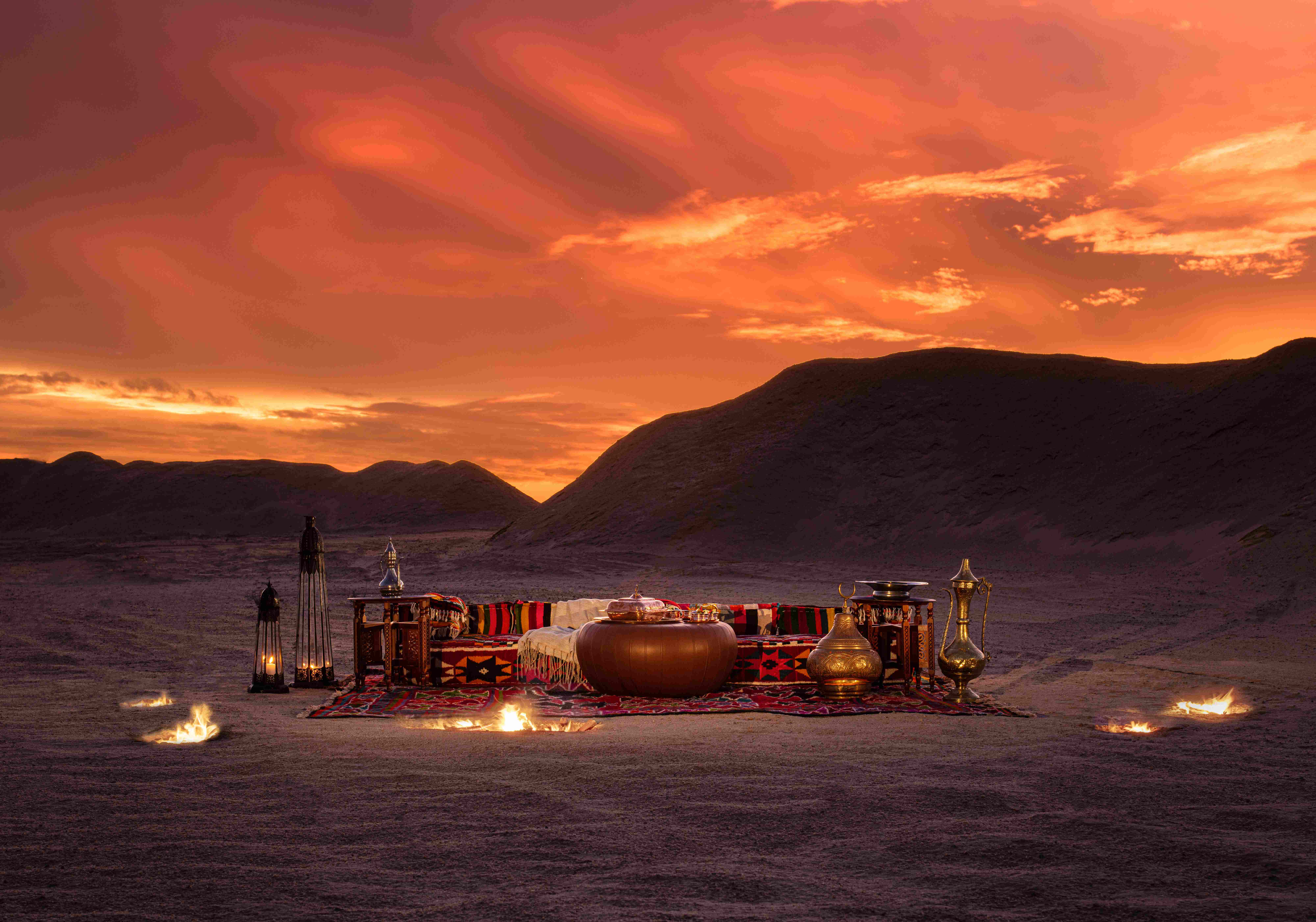 Romantic desert dinner setup at golden sunset with traditional Moroccan seating featuring red and black patterned cushions, brass lanterns, illuminated fire bowls, sand dunes, and dramatic fiery orange sky