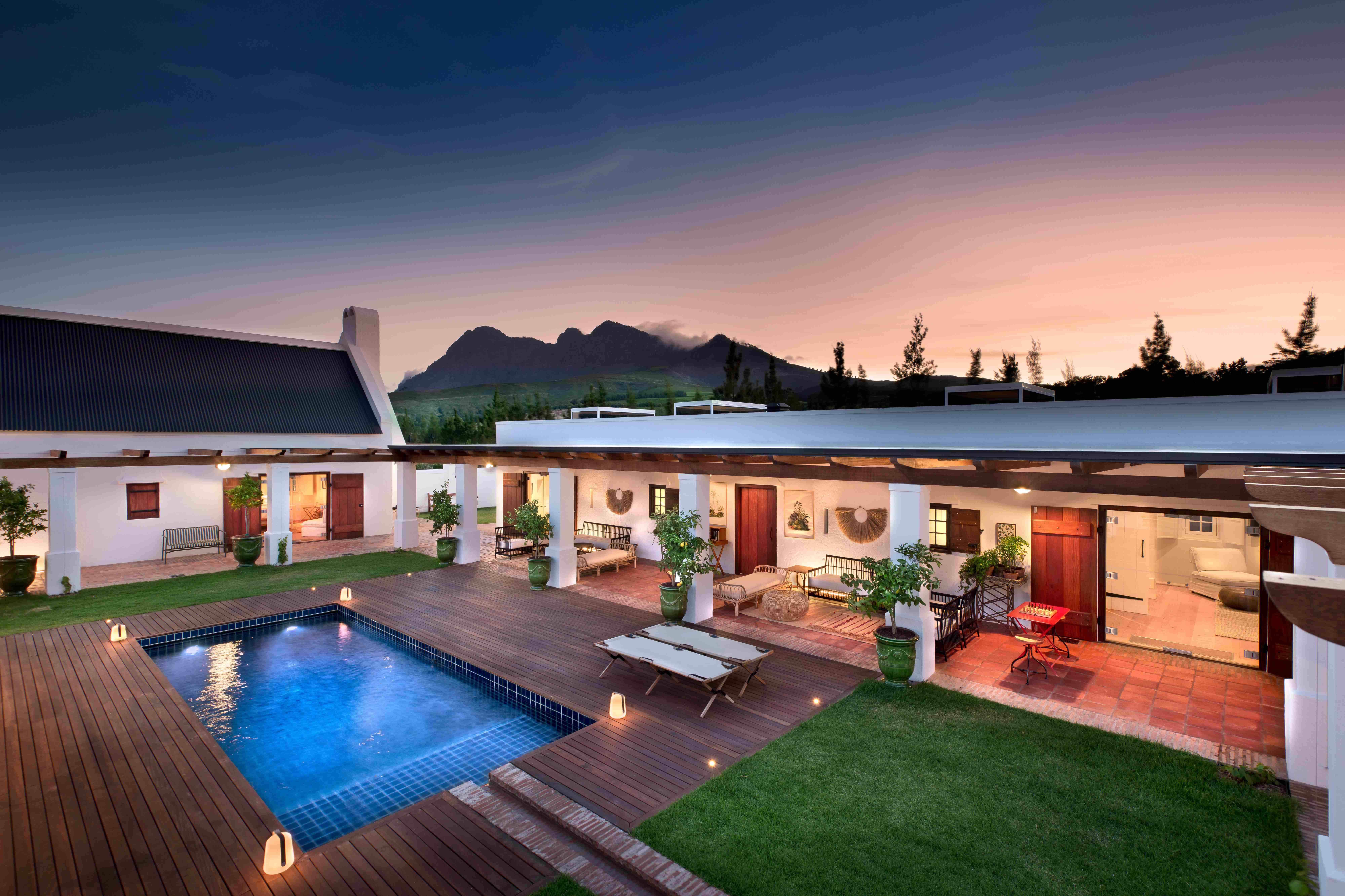 Expansive Fynbos Family House at twilight with illuminated white architecture, dark timber deck surrounding a sapphire-blue heated pool with uplighting, white columns supporting verandah, indoor/outdoor living spaces, lounge chairs, potted plants, manicured green lawn, and dramatic mountain backdrop under evening sky