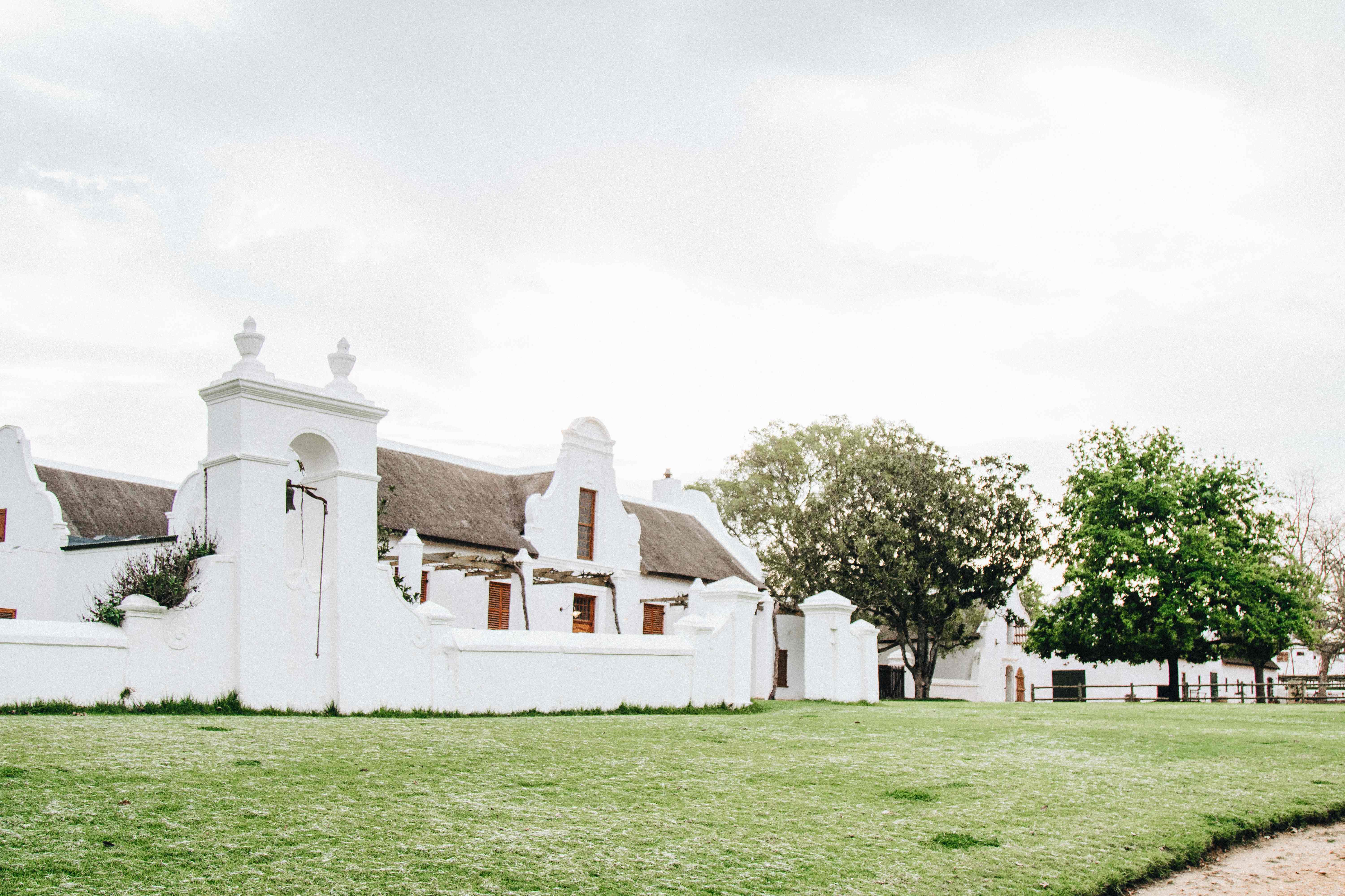 Charming Koornhuis Cape Dutch building with pristine white-washed walls, multiple thatched roof sections, bell tower with finial, brown wooden doors and windows, set on expansive green lawn with mature native trees under clear sky, embodying traditional Winelands architecture