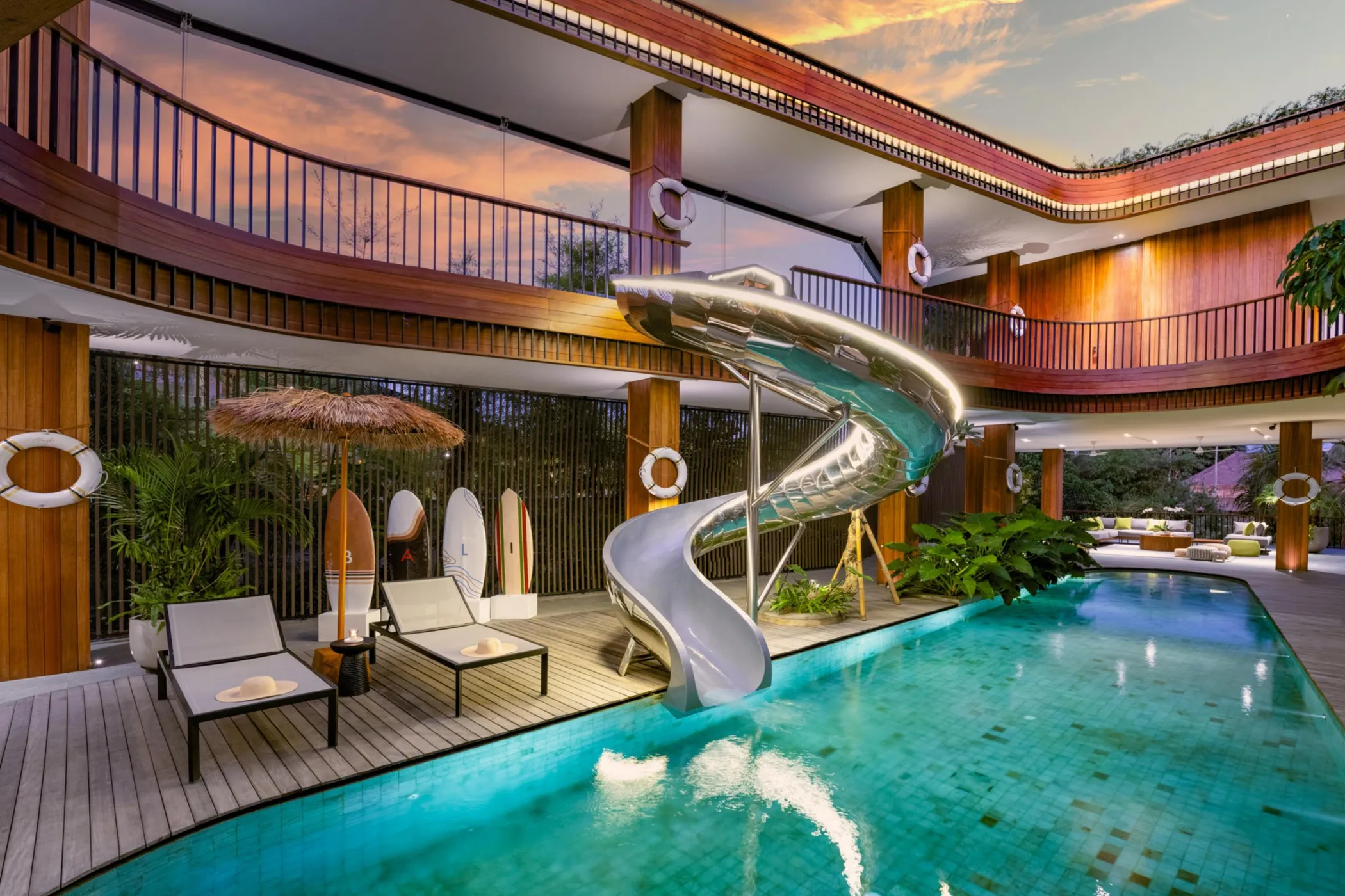 Luxurious modern Akashi Residence interior featuring a distinctive silver spiral water slide ending in an emerald-turquoise pool, lounge chairs, wooden deck, tropical plants, and dramatic architecture with warm golden evening lighting