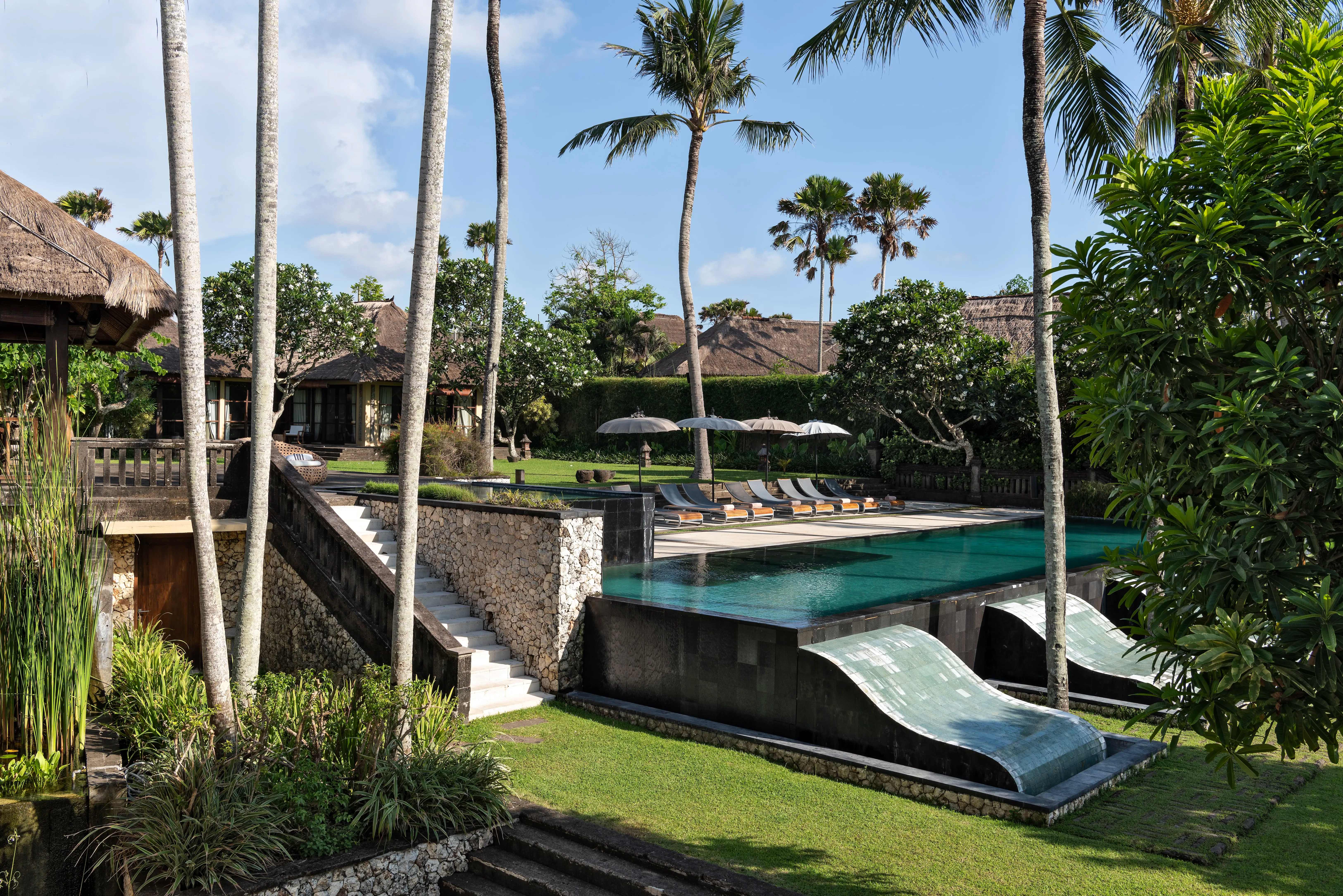 Traditional Balinese Villa Mary with thatched-roof pavilions, lush tropical gardens with tall coconut palms, turquoise pool with waterslide, emerald vegetation, and authentic tropical estate ambiance