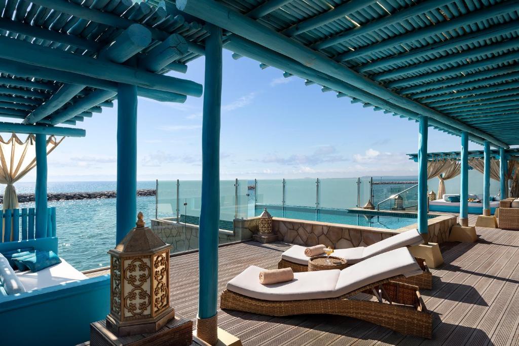 Modern luxury island resort terrace with turquoise architectural columns and pergola, white cushioned loungers, brass decorative lanterns, ocean views, and glass railings