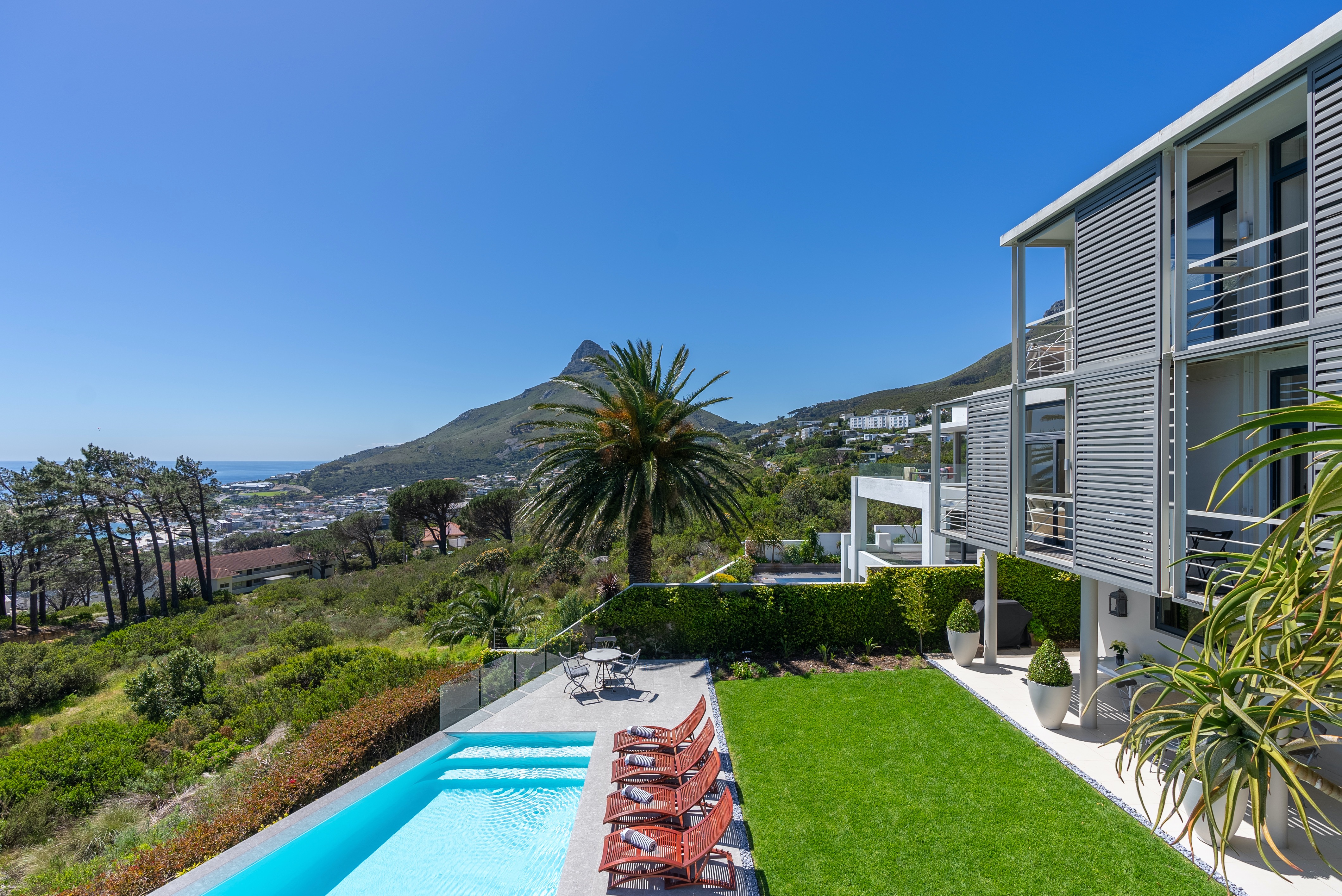 Contemporary villa with white and gray tones featuring turquoise lap pool, red loungers, manicured lawn, palm trees, and panoramic views of mountains and Atlantic coastline