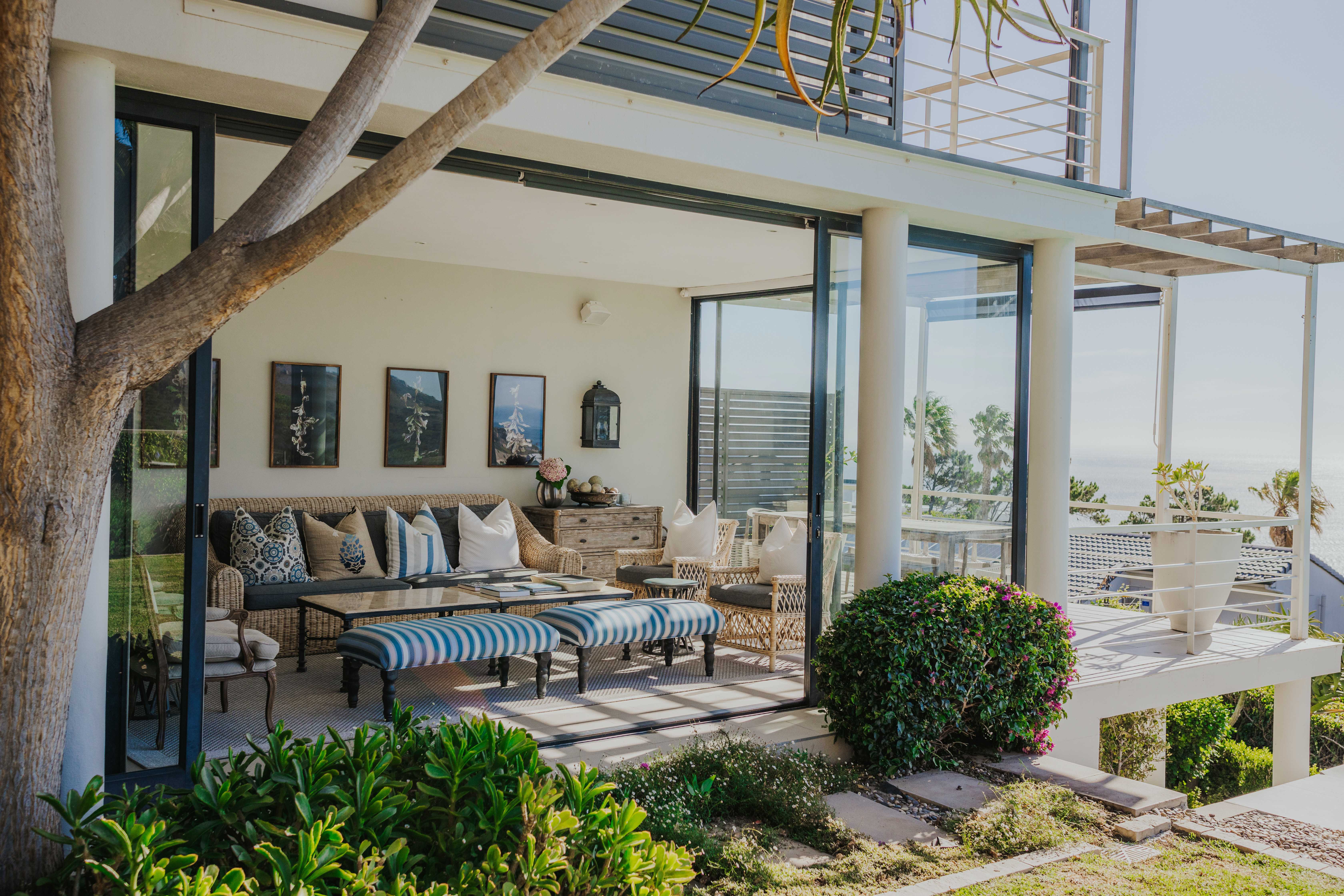 Indoor-outdoor living space with covered patio, rattan and striped coastal seating, woven furniture, glass doors opening to ocean views, and seamless interior-exterior flow