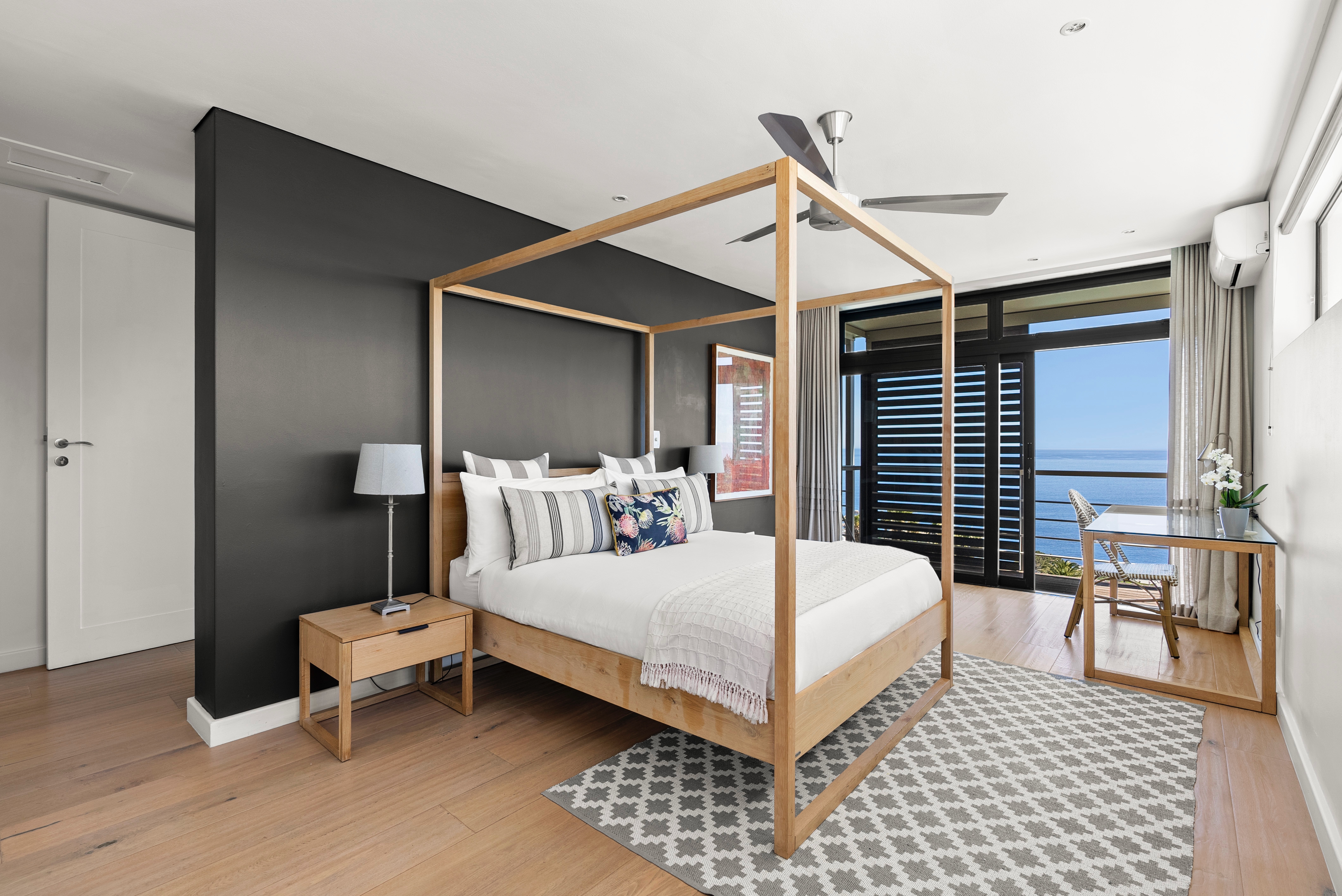 Luxurious master bedroom with wooden four-poster bed, black accent wall, white bedding with patterned pillows, large ocean-view doors, cream curtains, and coastal contemporary design
