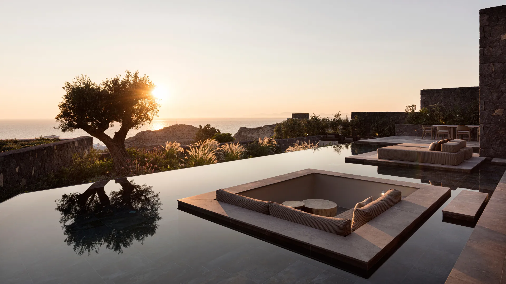 Luxury villa with infinity pool edge, black stone elements, lounge seating, and Aegean Sea sunset views