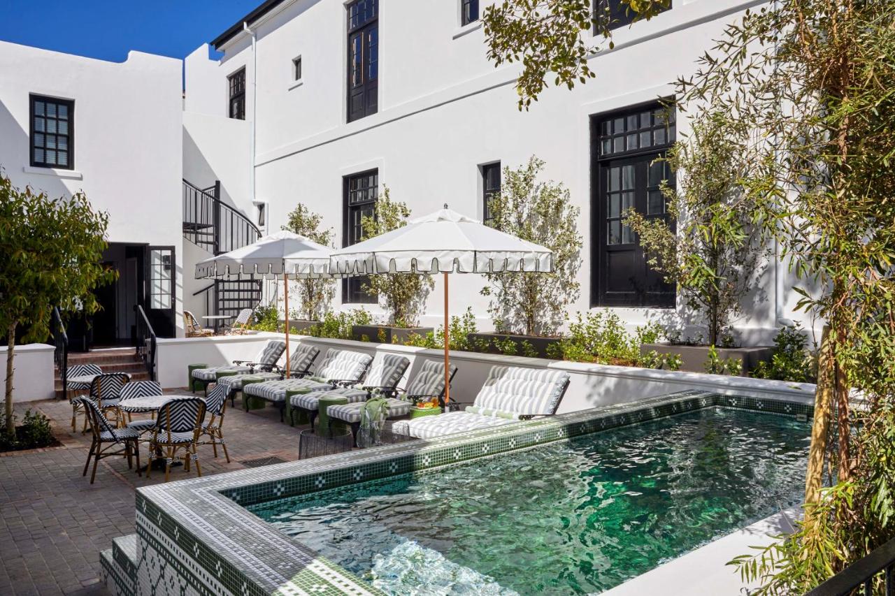 Emerald green swimming pool with decorative tile details, white umbrella-covered loungers with striped cushions, colonial white building facade with black trim, climbing ivy, paved terrace, bright daylight