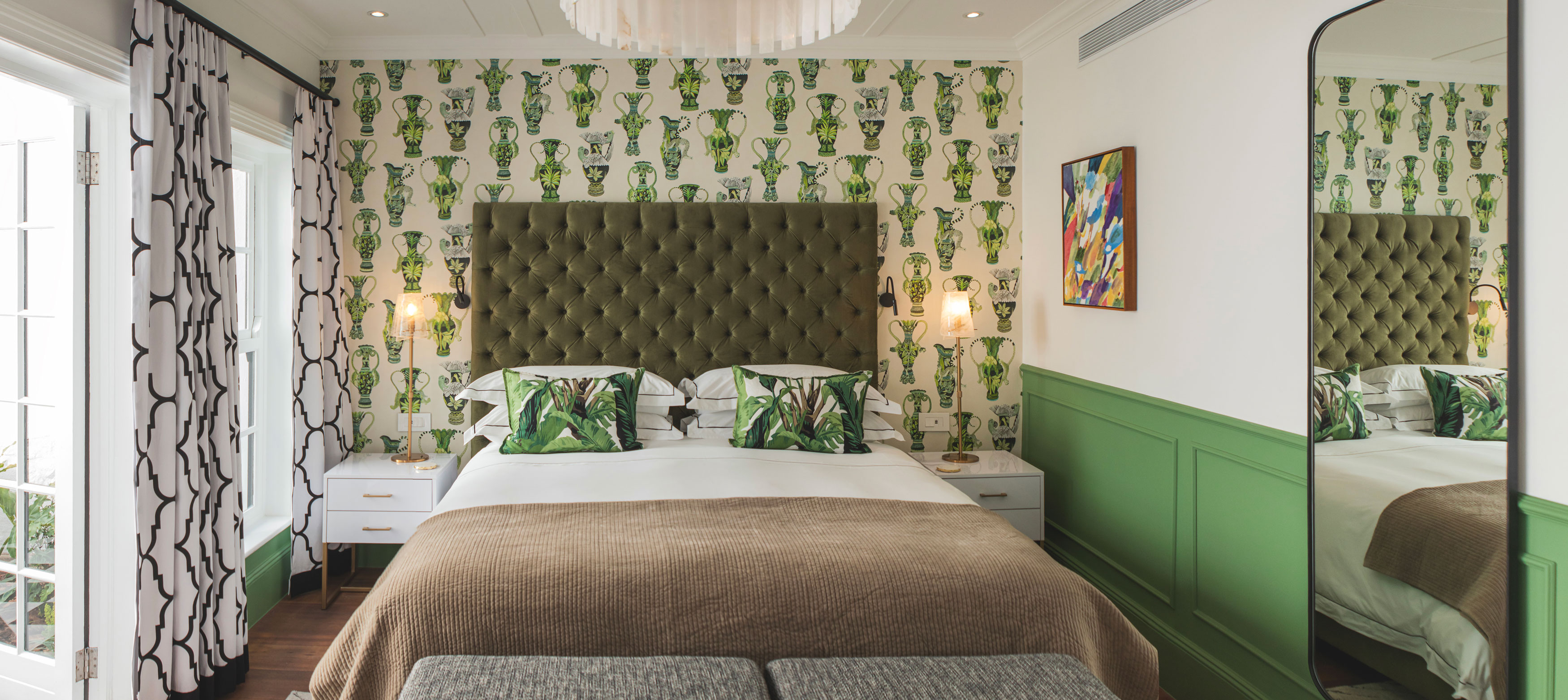 Sophisticated bedroom interior with green botanical wallpaper, olive-green quilted headboard, white bedside tables with table lamps, green patterned curtains, large mirror, botanical throw pillows, warm interior lighting