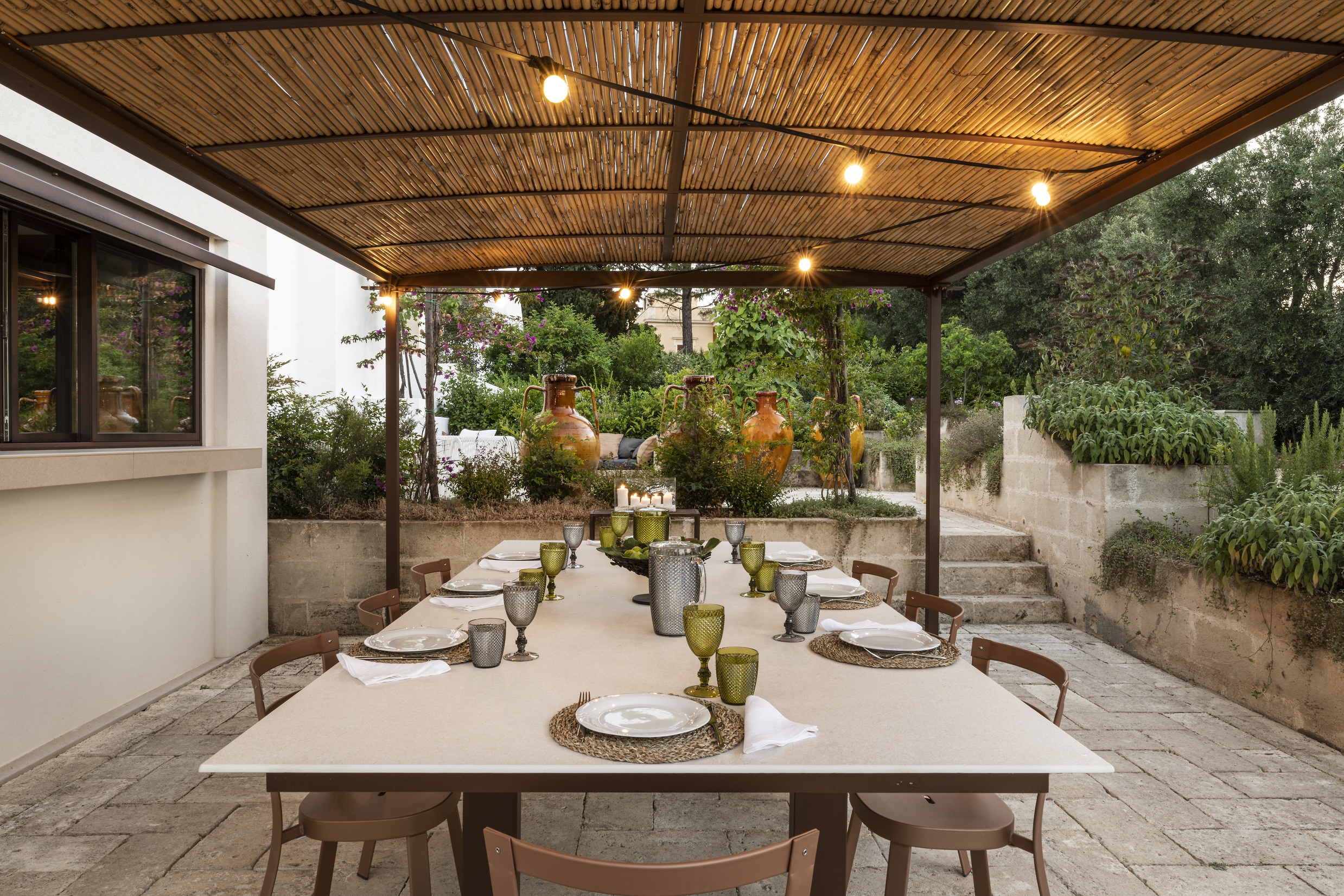 Elegant covered dining terrace with wooden pergola, warm string lighting, table set with golden vessels and green glassware, stone walls, Mediterranean gardens