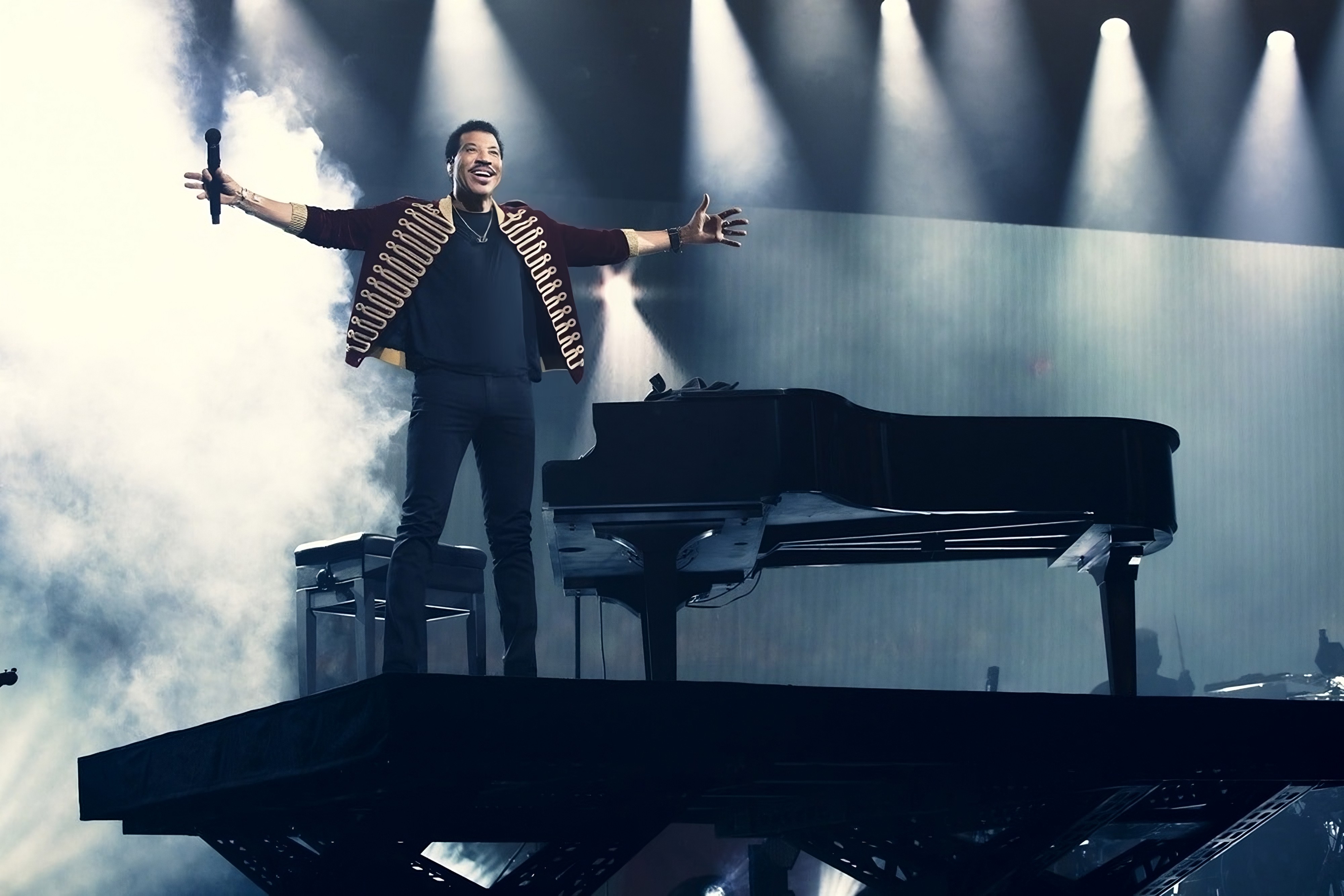 Lionel Richie performing on stage with arms spread wide wearing a decorated jacket with grand piano