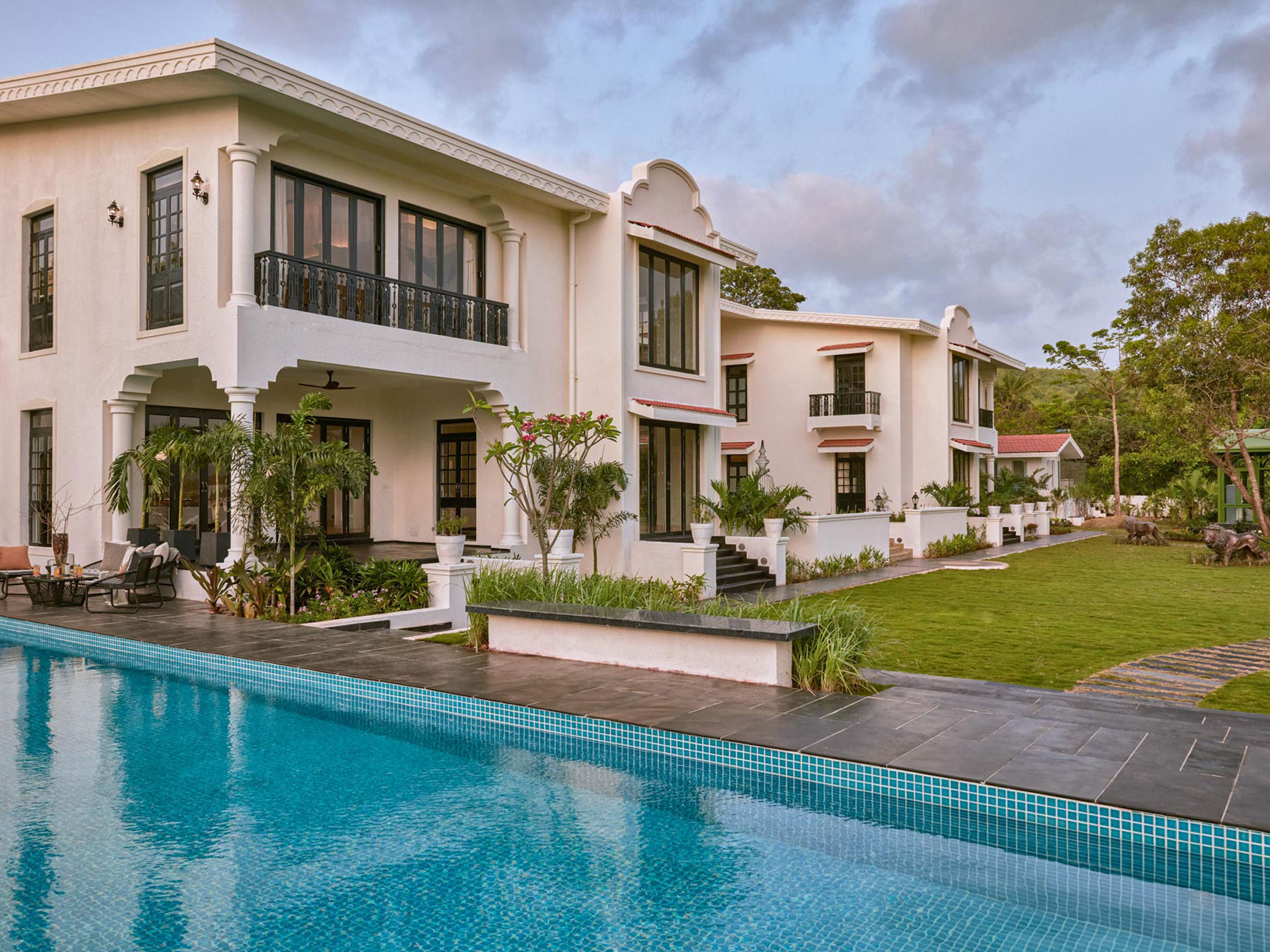 Charming multi-story colonial villa in cream and pale pink with black-framed windows, red architectural accents, turquoise tile-lined pool, manicured green lawn, tropical plantings with frangipani trees, and traditional Indian colonial design aesthetic