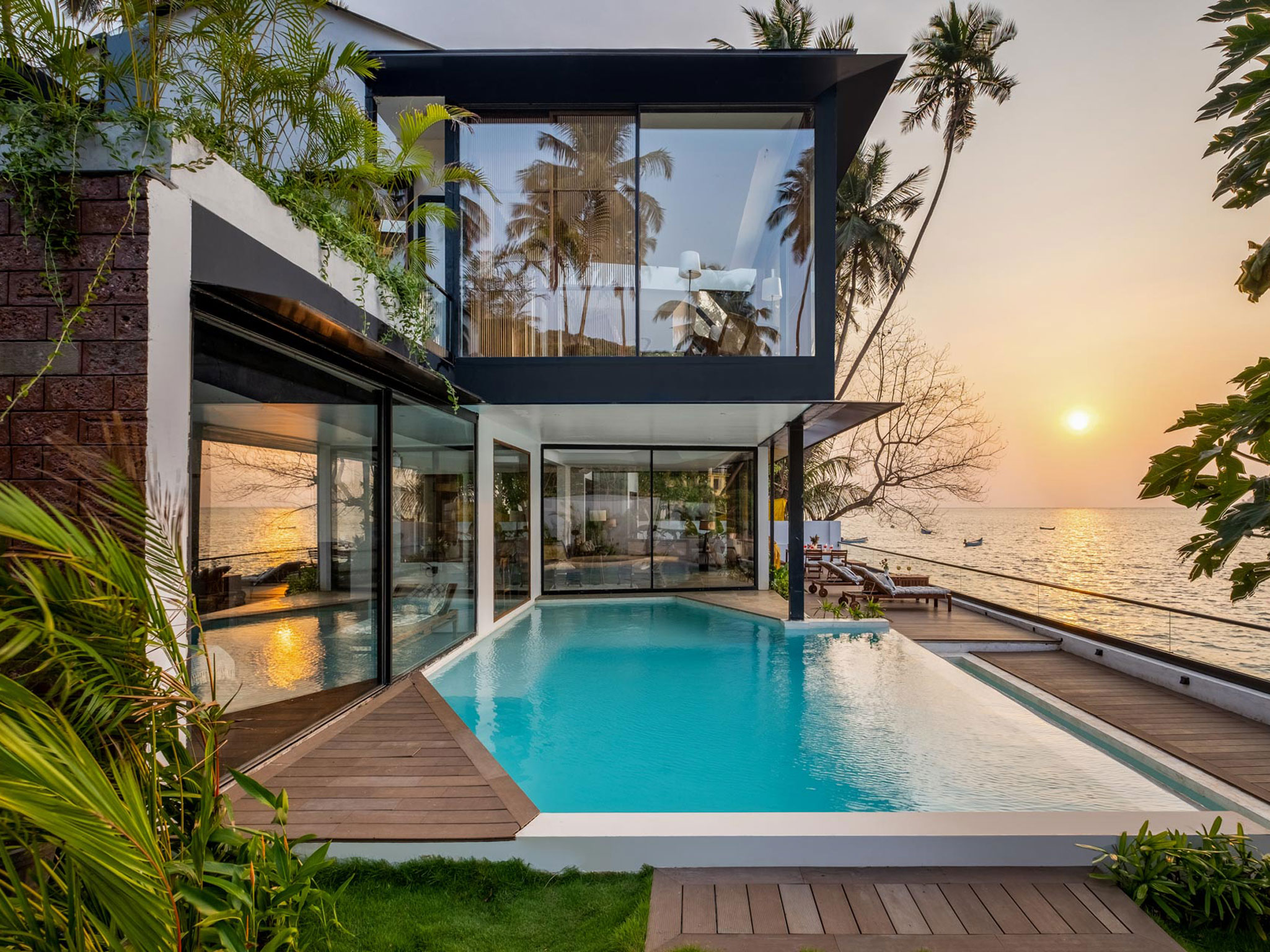 Contemporary luxury beachfront villa at golden hour with modern black-framed glass architecture, turquoise infinity pool, wooden deck terrace, palm tree landscaping, white interior visible through expansive windows, ocean sunset view, and minimalist design