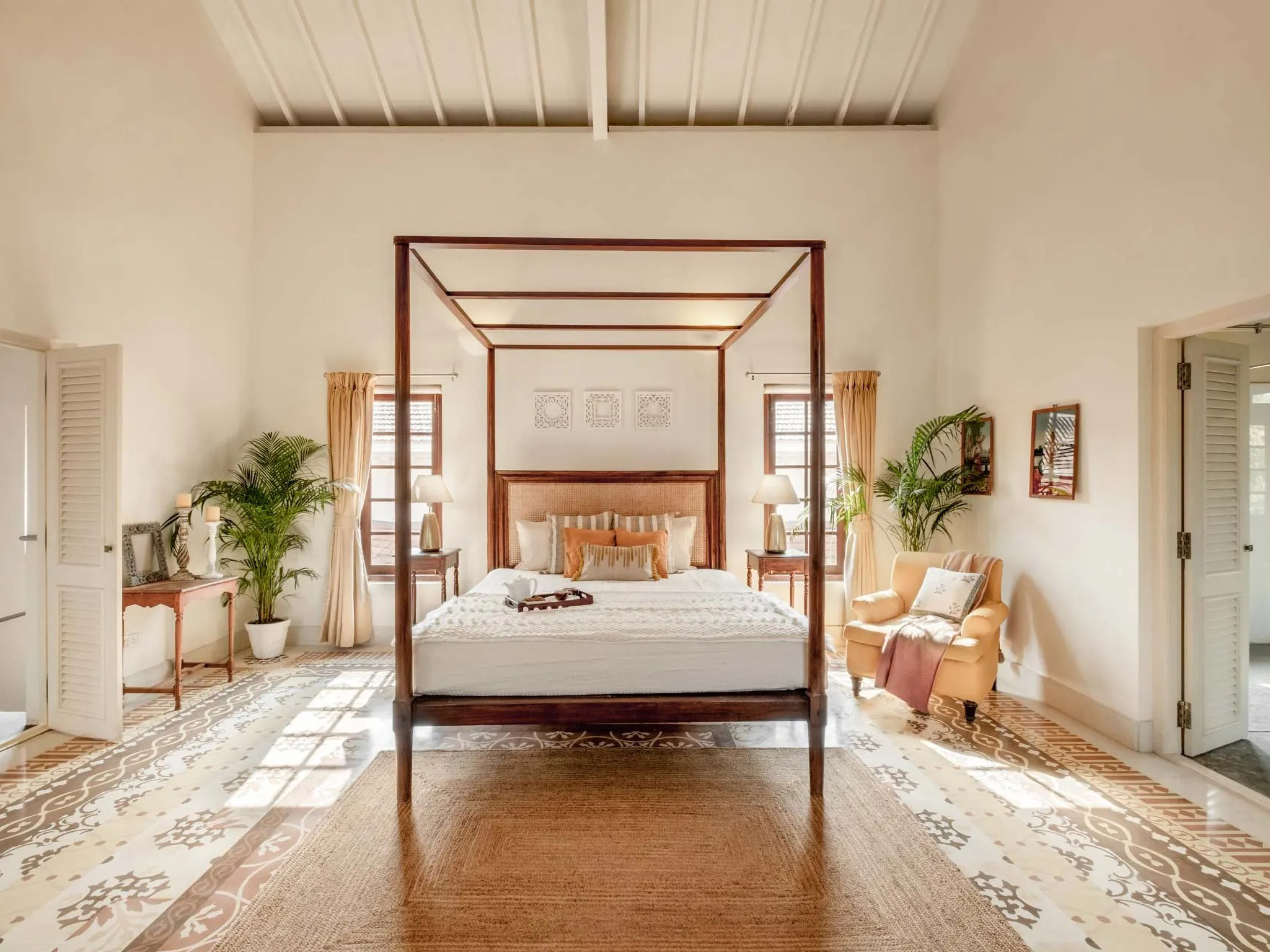 Elegant master bedroom suite with four-poster canopy bed in warm reddish-brown wood, cream and gold bedding, white ceiling with architectural beams, potted tropical plants, traditional patterned flooring, wooden furniture pieces, and shuttered windows creating serene colonial-inspired luxury