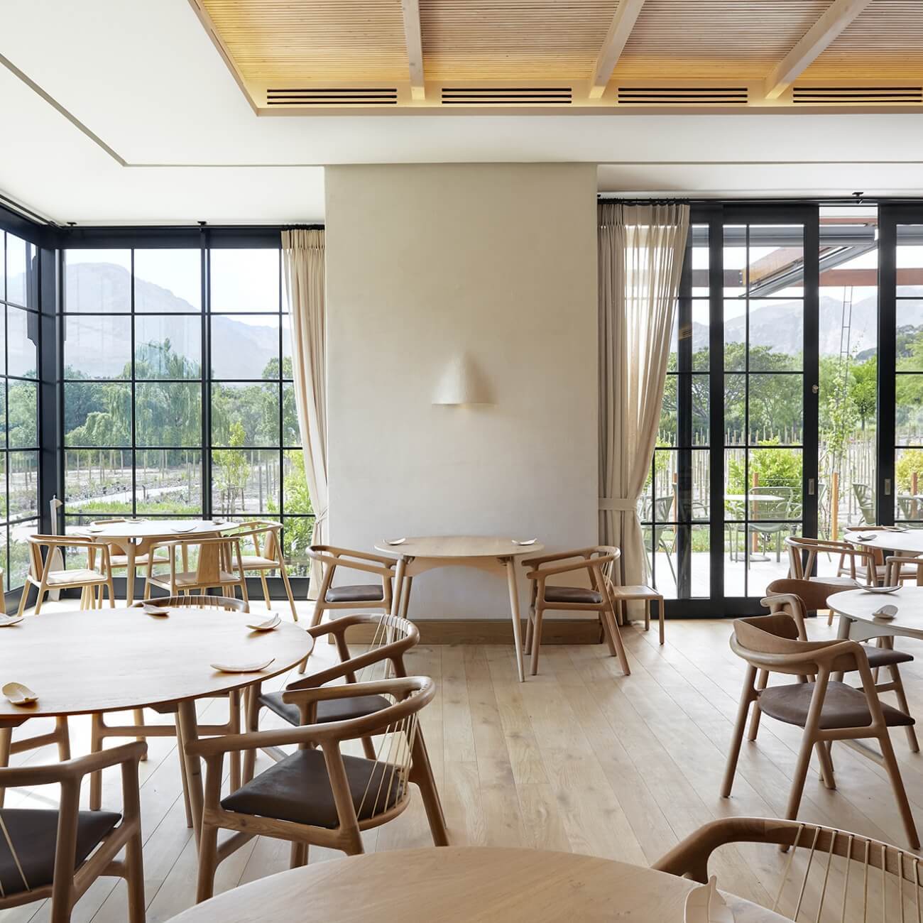 Modern fine dining restaurant with floor-to-ceiling windows overlooking vineyard mountains, light wood furniture and contemporary design