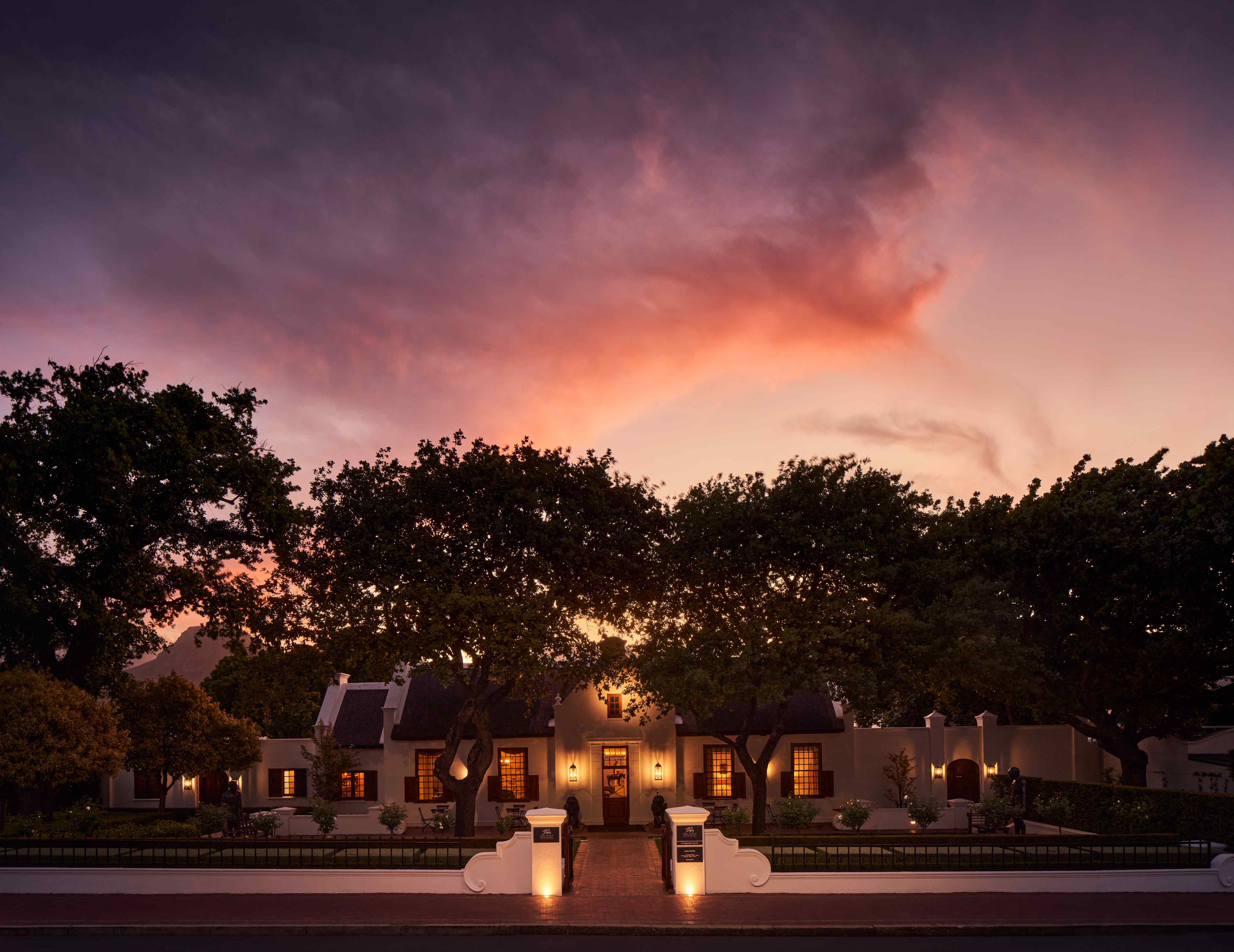 Luxury estate at dusk with Cape Dutch architecture, illuminated windows, dramatic sunset sky and ancient oak trees