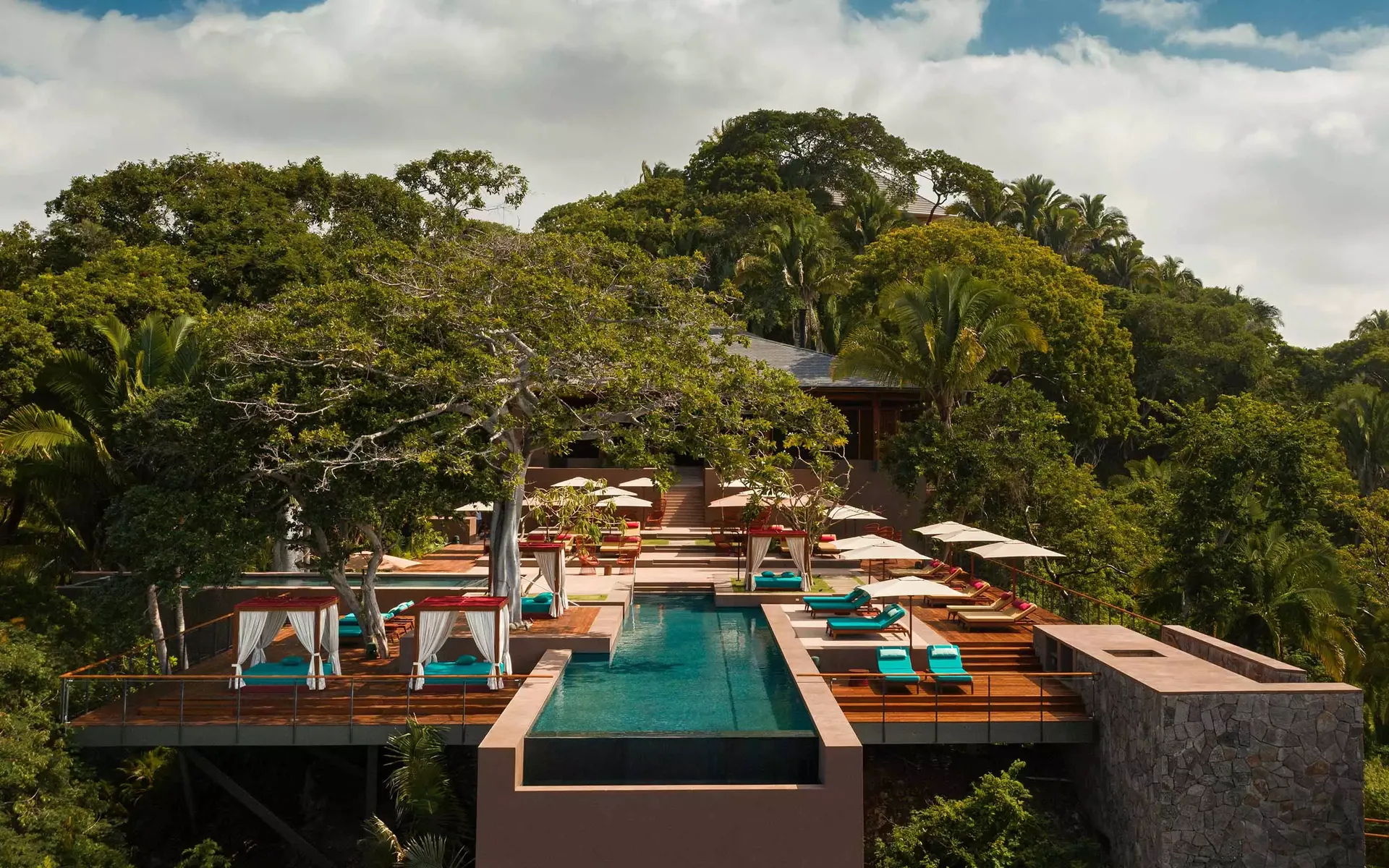 Aerial view of luxury resort villa with long turquoise infinity pool, turquoise-cushioned lounge areas, white umbrellas and cabanas, burnt orange terraces, dense surrounding jungle landscape