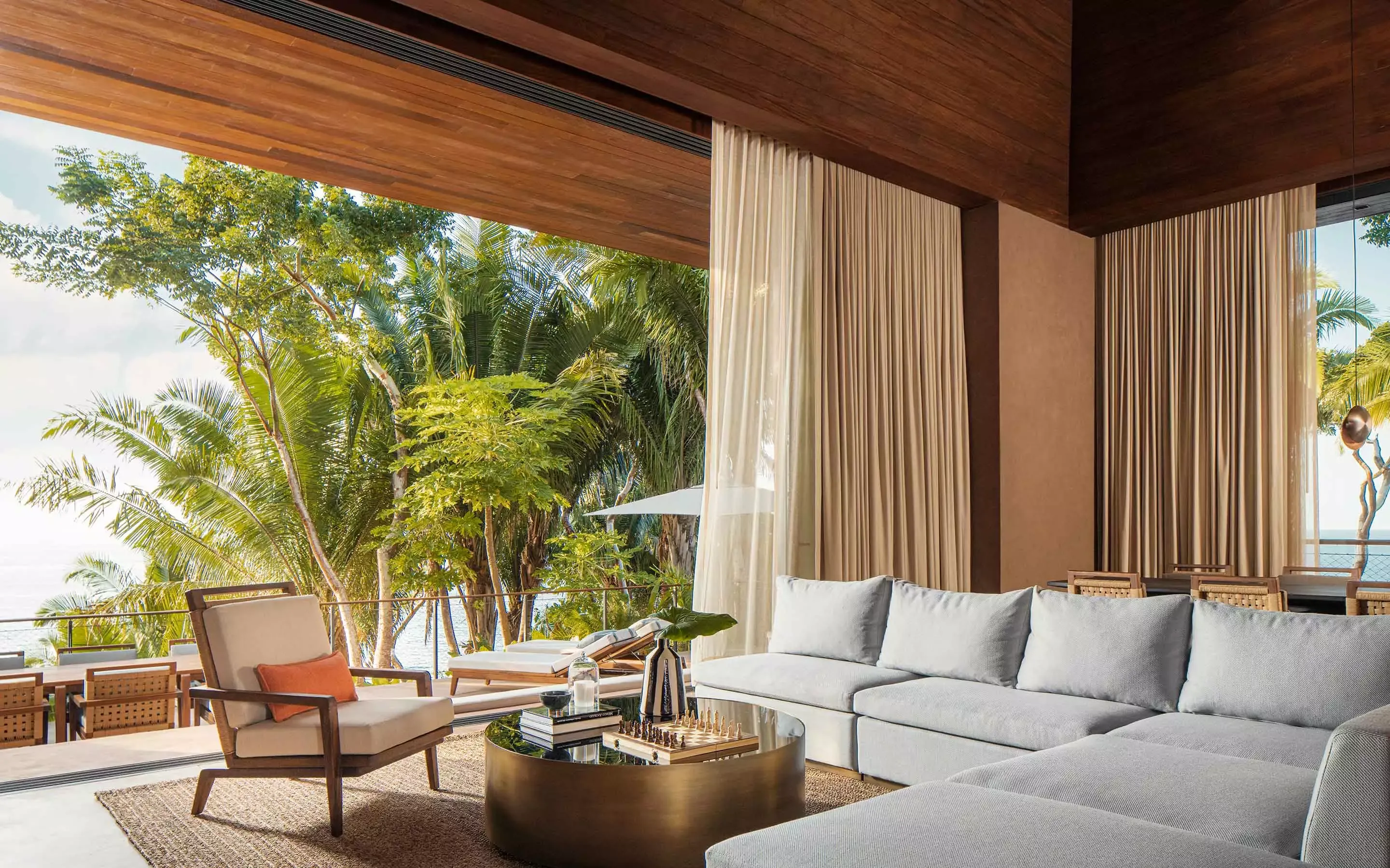 Modern villa interior living space with warm wood ceiling and wall treatments, cream sofa with orange accent pillow, metal base coffee table, wooden armchair, cream drapes, tropical ocean and palm tree views through large windows