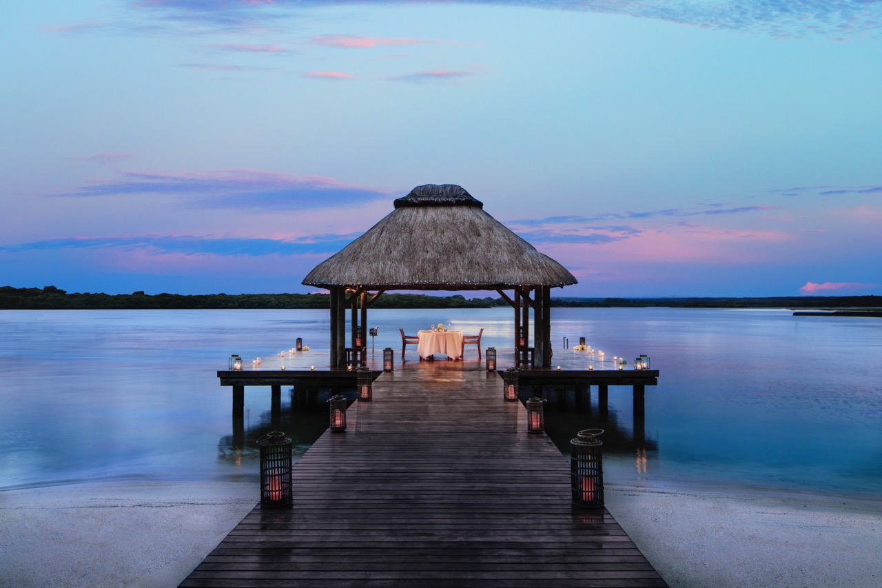 Romantic candlelit dinner pavilion at sunset over lagoon with thatched-roof gazebo and glowing lanterns