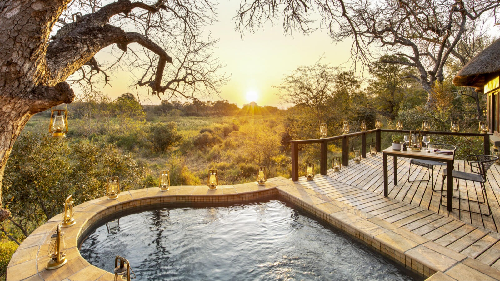 Infinity pool overlooking golden valley landscape with wooden dining table set for dinner, brass lanterns, gnarled acacia tree, sunset lighting