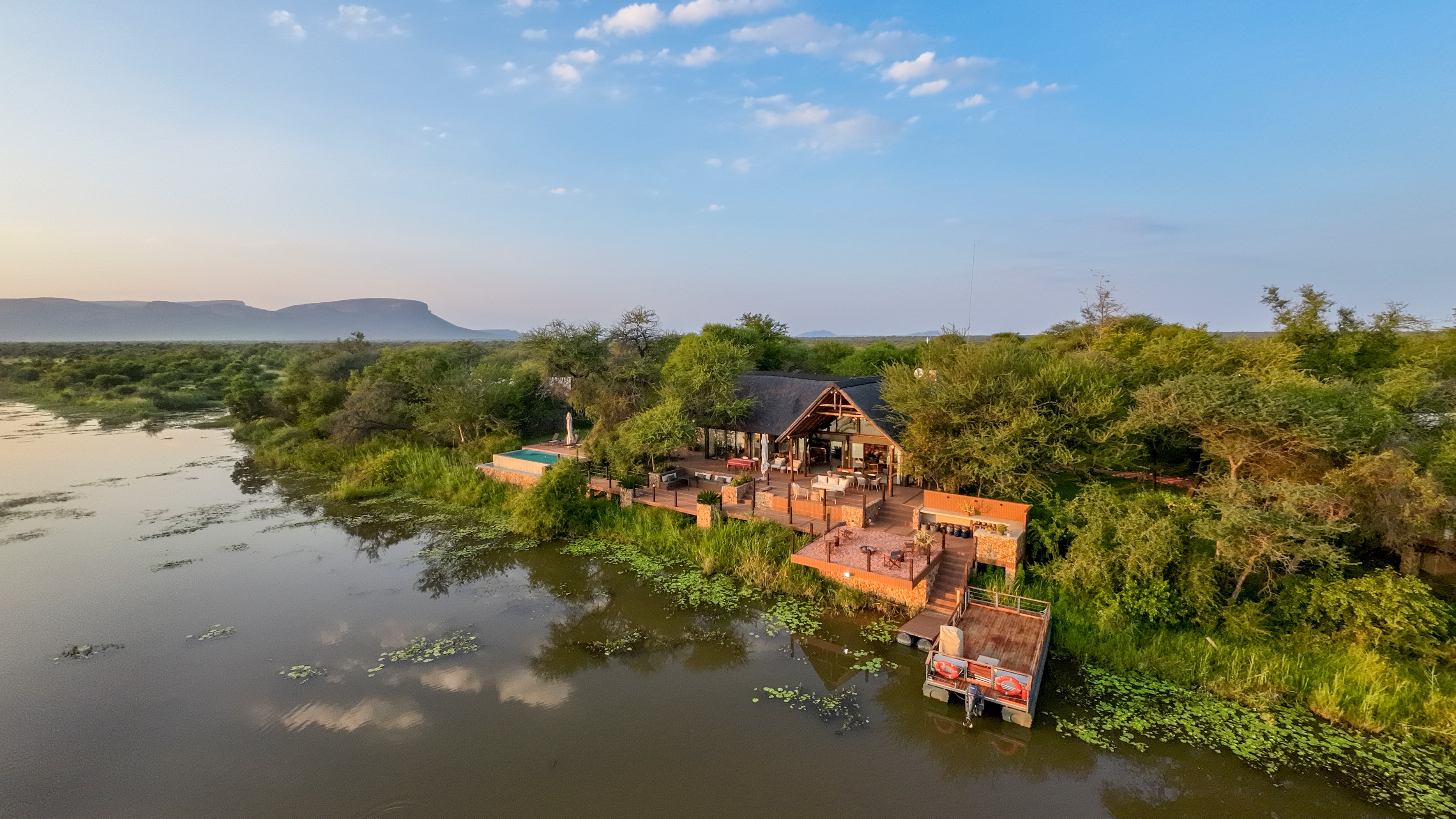 Aerial bird's-eye view of riverside lodge complex with thatched structures, turquoise pool, wooden decks, boat dock, lush green bushveld vegetation, mountains on horizon