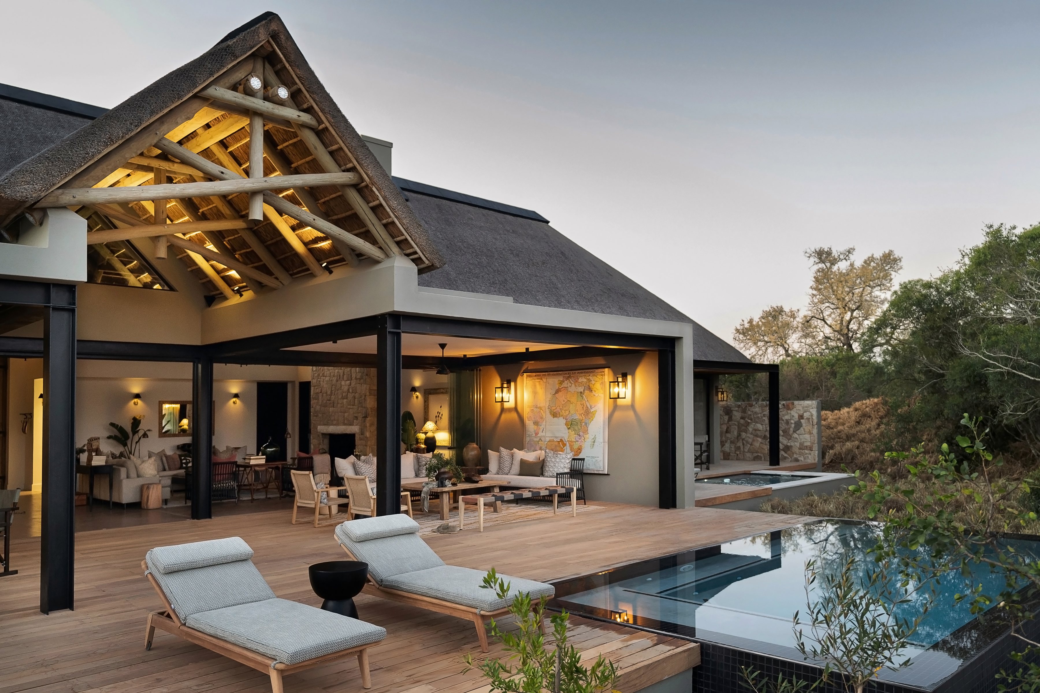 Modern luxury lodge with illuminated thatch roof structure, wooden deck with cushioned lounge chairs, infinity pool edge, stone walls, glass doors, bushveld landscape backdrop