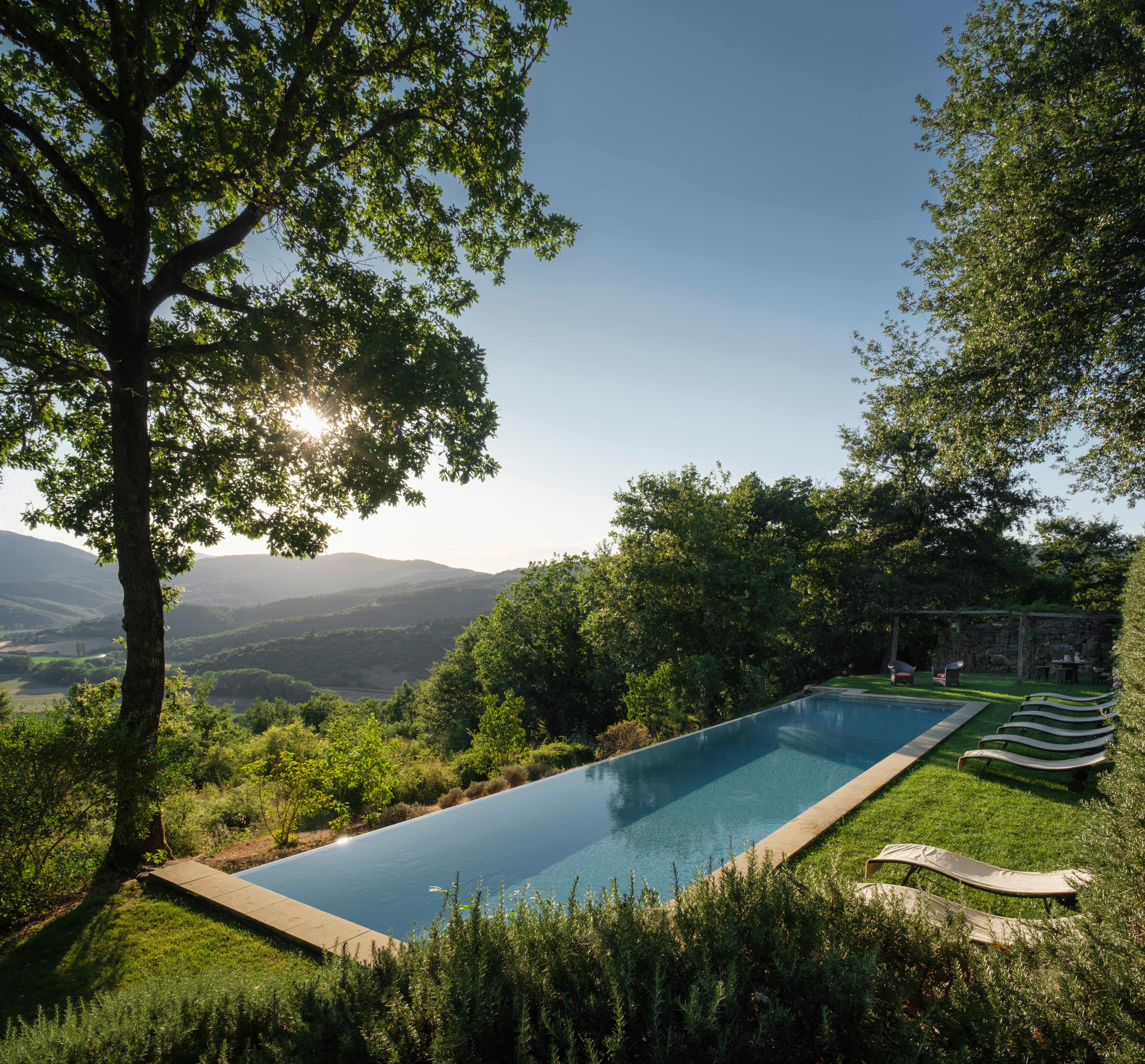 Infinity pool overlooking Umbrian valley with mountains in distance, loungers beside pool, tall trees providing shade, lush green landscaping