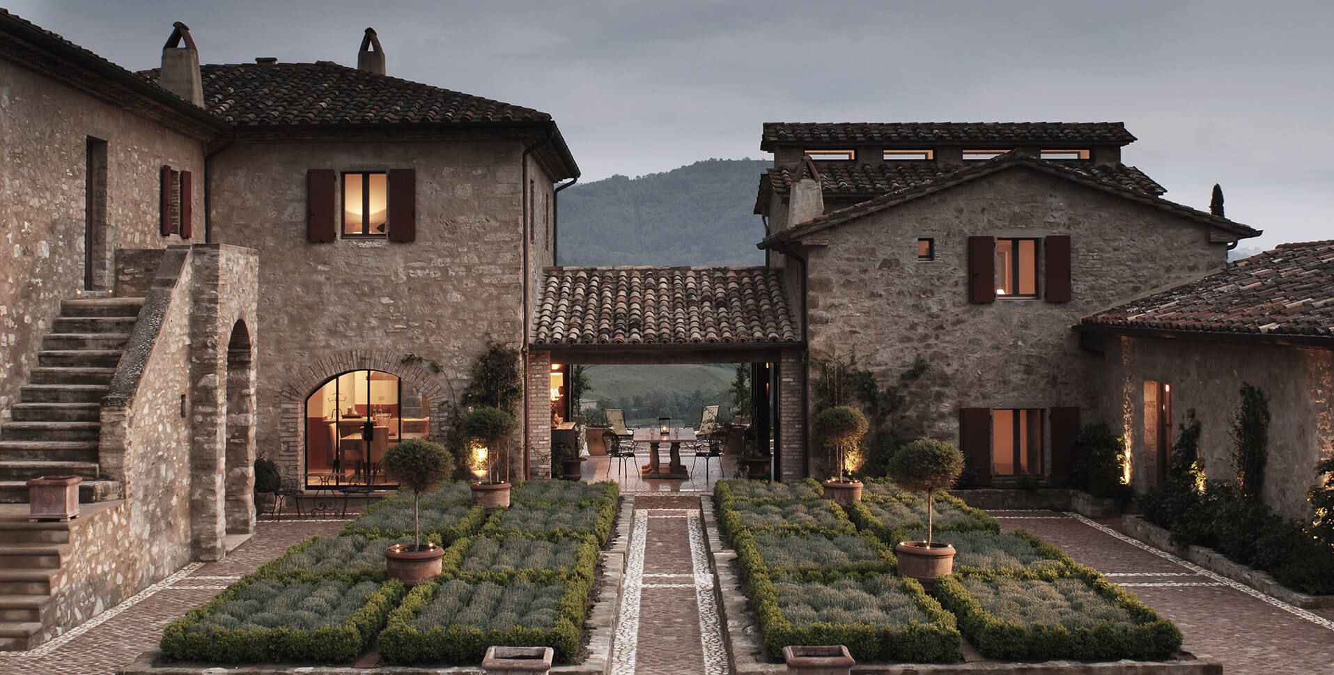 Traditional Italian stone farmhouse courtyard at twilight with illuminated windows, terracotta tiled roofs, lavender hedges lining central pathway, warm interior lighting, mountains in background