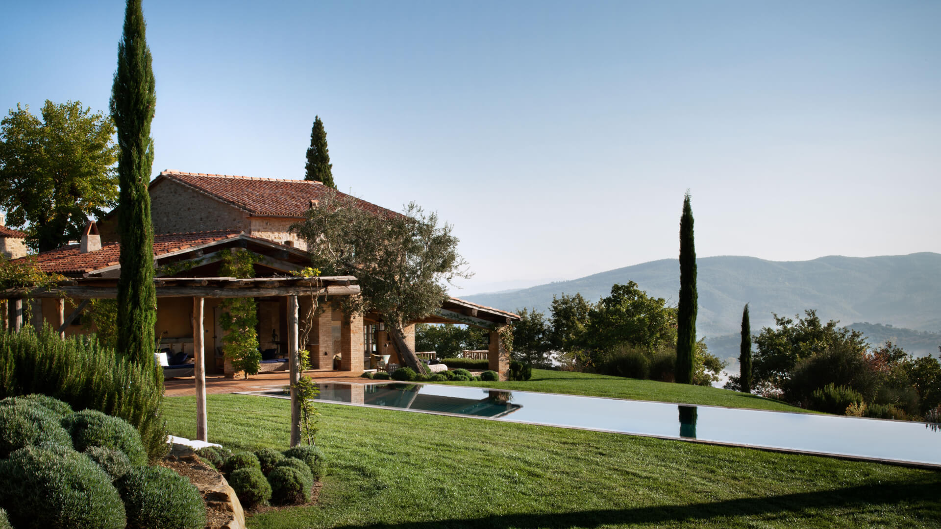 Rustic Tuscan-style villa with terracotta tile roof, ochre stone walls, wooden pergola, infinity pool overlooking valley, cypress trees, manicured green lawn, rolling hills and mountains