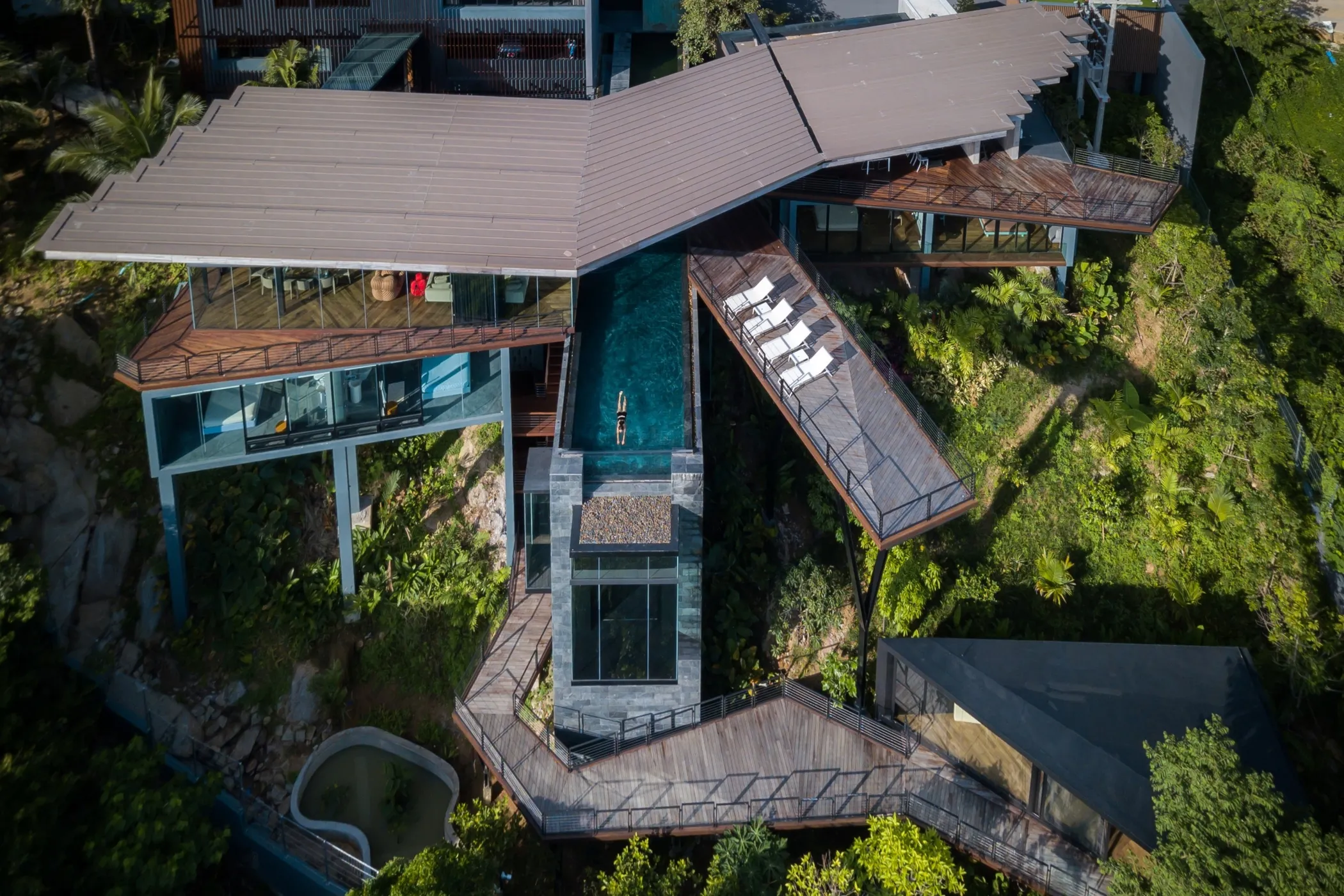 Aerial drone view of modern Kelapa House with multiple terraced levels, turquoise 17-meter infinity pool, wood and white contemporary materials, surrounded by lush tropical vegetation on Koh Samui hillside