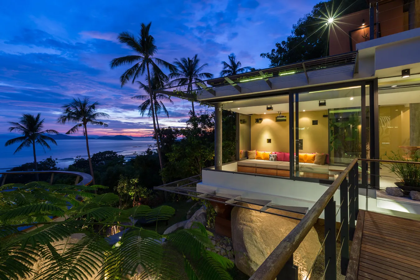 Twilight view of L2 Residence with minimalist glass-fronted modern pavilion, warm interior glow, white infinity pool edge, tropical vegetation, ocean and island views in background under dramatic sunset sky