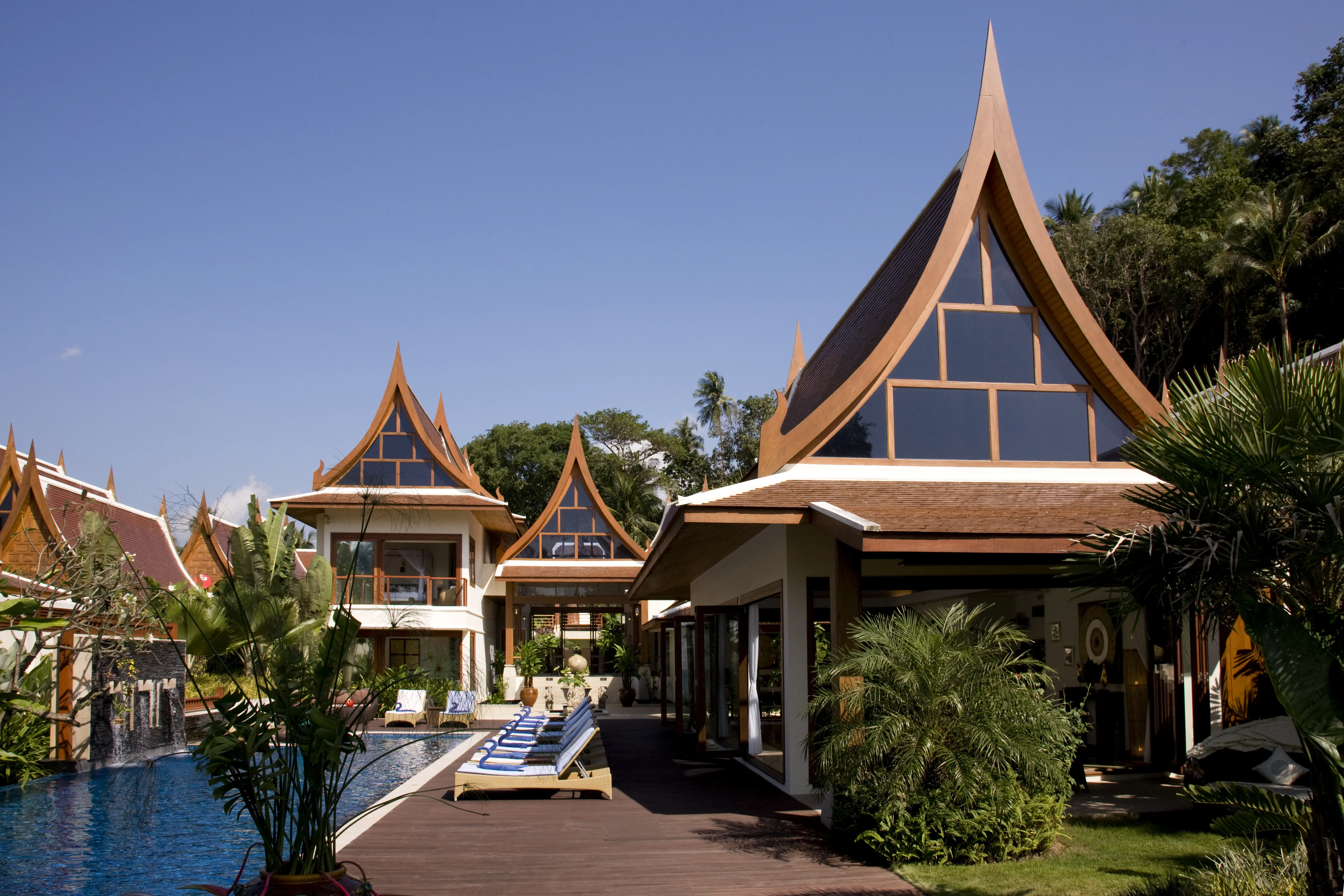 Villa Haineu showcasing distinctive Thai architecture with peaked golden/tan roofs reminiscent of traditional temples, modern white base, blue infinity pool with loungers, tropical palms and foliage, clear sky