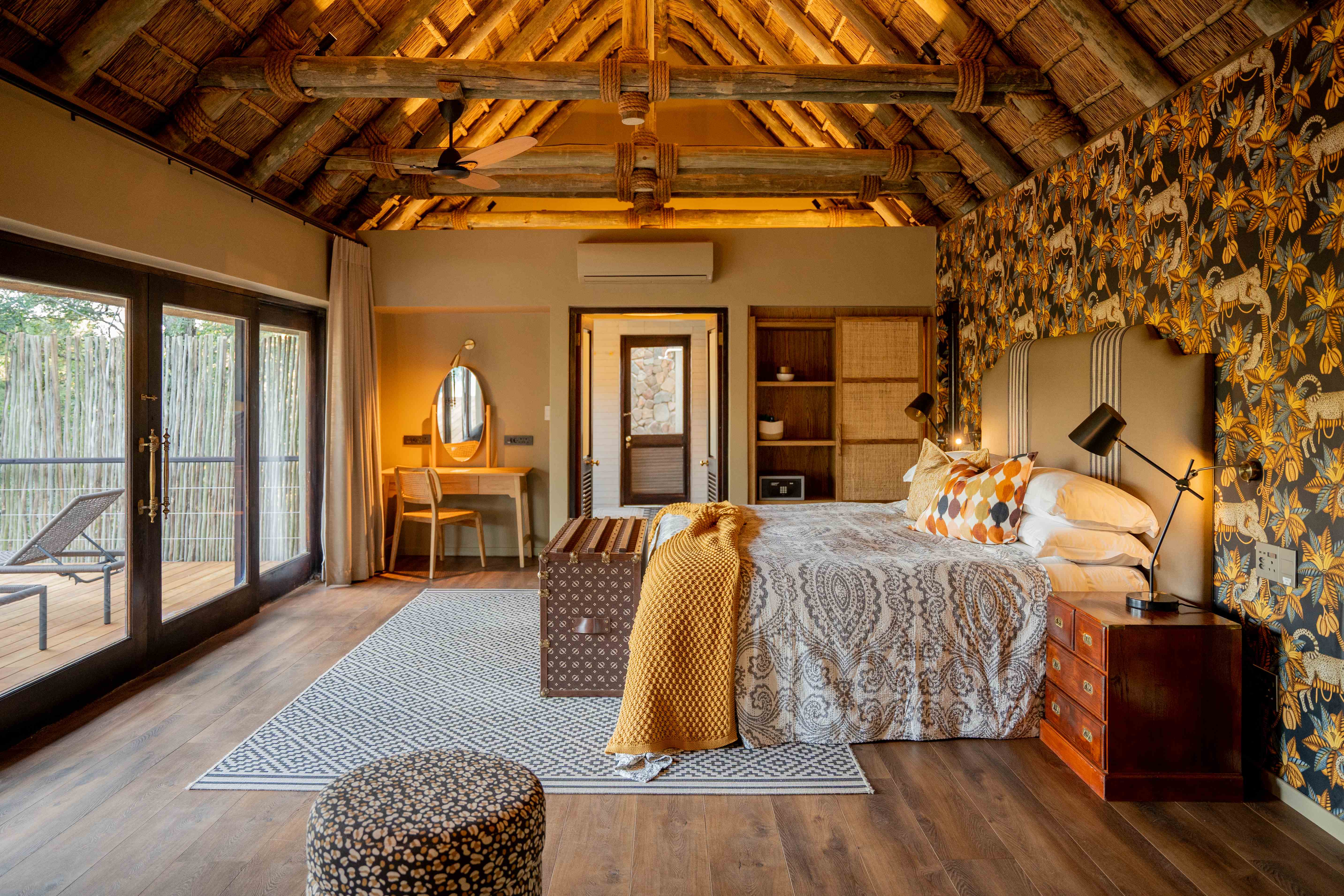 Luxurious bedroom with golden tones, exposed wooden ceiling beams, yellow patterned wallpaper, king bed with layered bedding, glass doors overlooking acacia trees