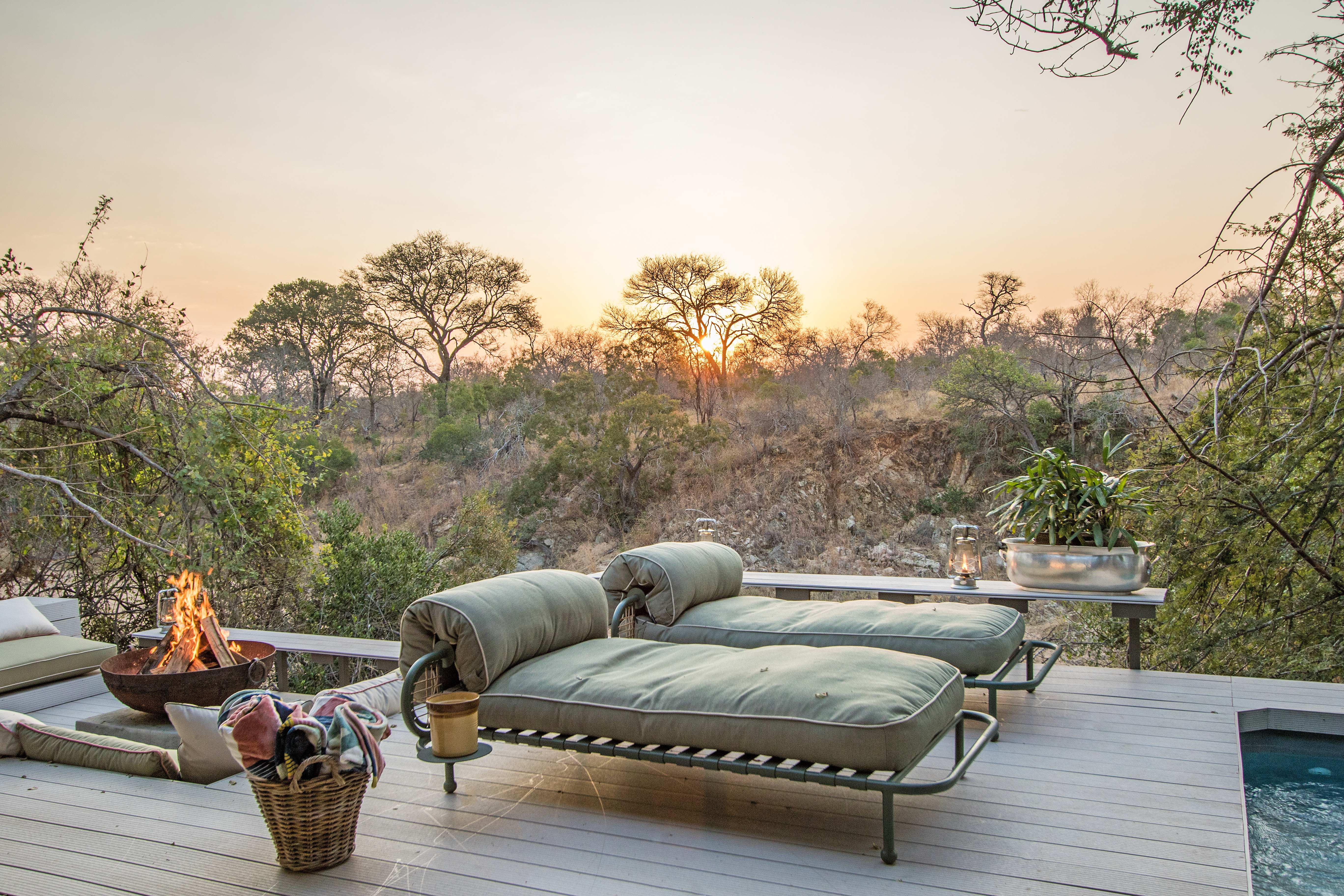 Sunset deck with fire pit, olive-green cushioned daybeds, wicker basket with blankets, overlooking bushveld valley with acacia trees, golden hour lighting