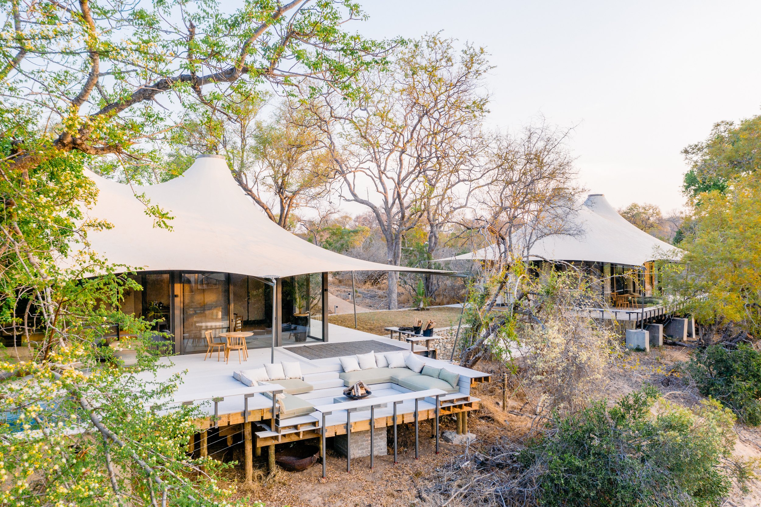Glamping pavilion with white canvas shade sails, cream cushioned sectional seating, wooden chairs, fire pit, thatched lodge structures in background, savanna landscape with acacia trees