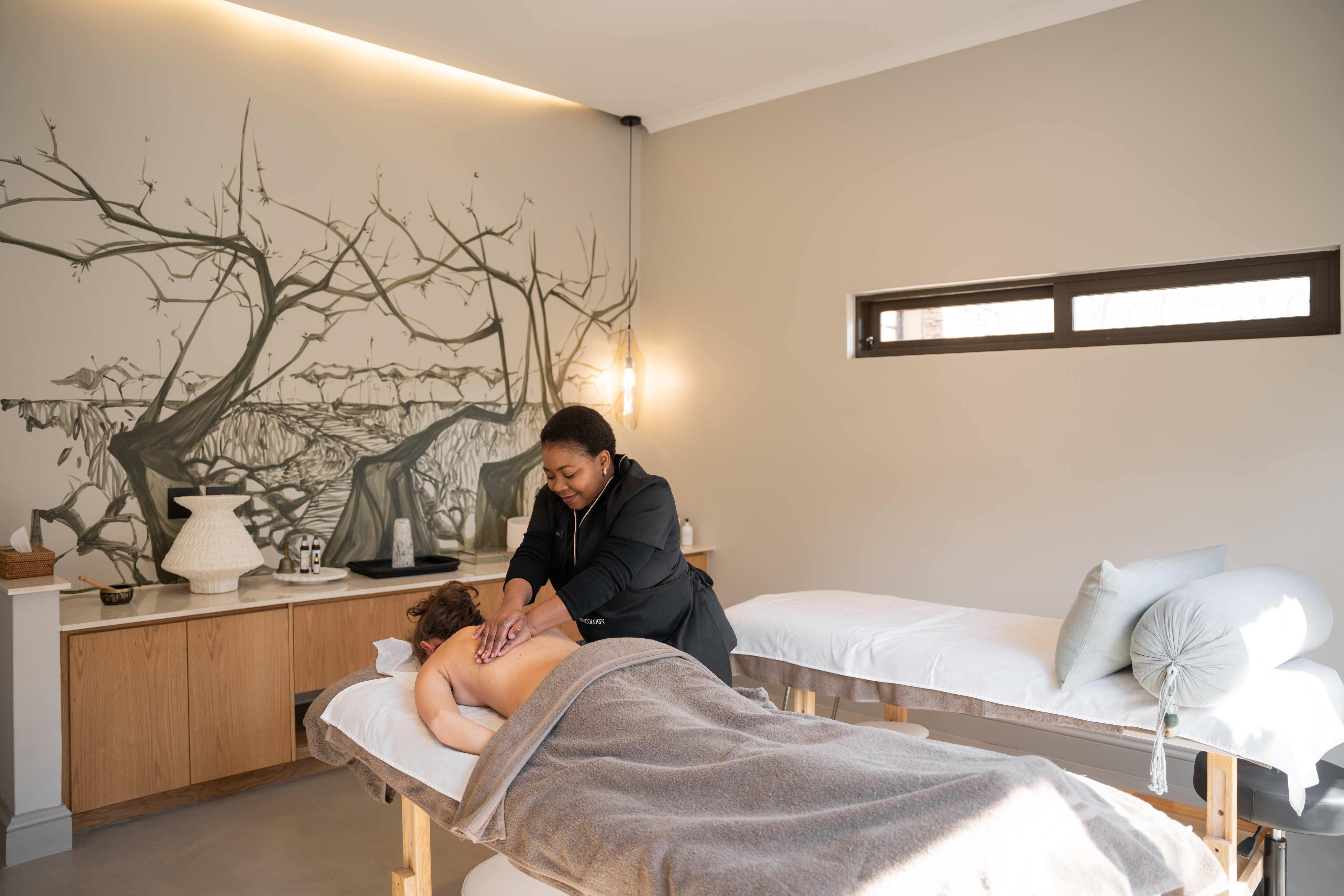 Modern spa treatment room with therapist performing massage, beige walls with artistic branch mural, white massage bed with pillows, wooden vanity with decorative items