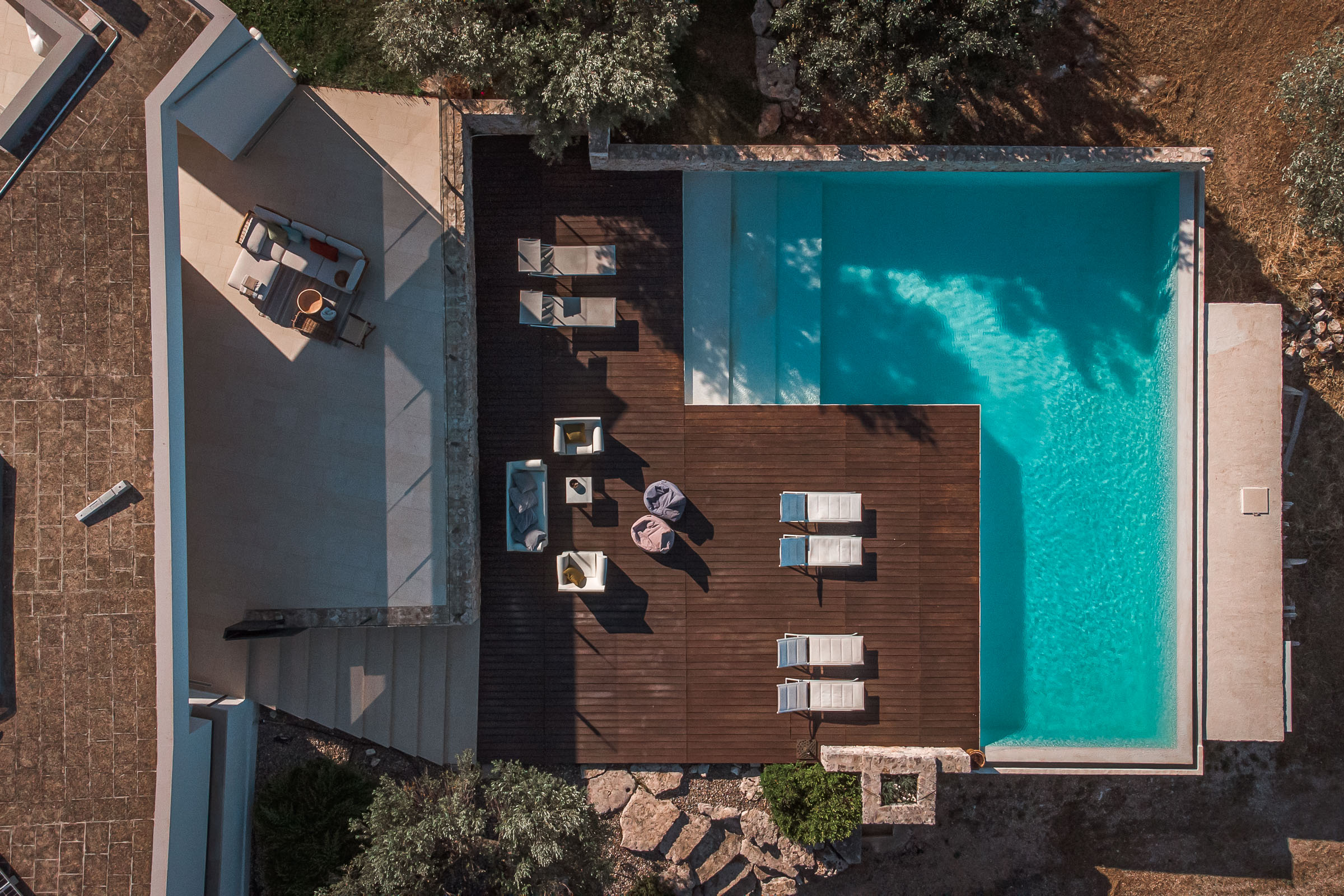 Aerial view of modern minimalist villa with teal infinity pool, wooden deck with loungers, contemporary architecture with Mediterranean vegetation