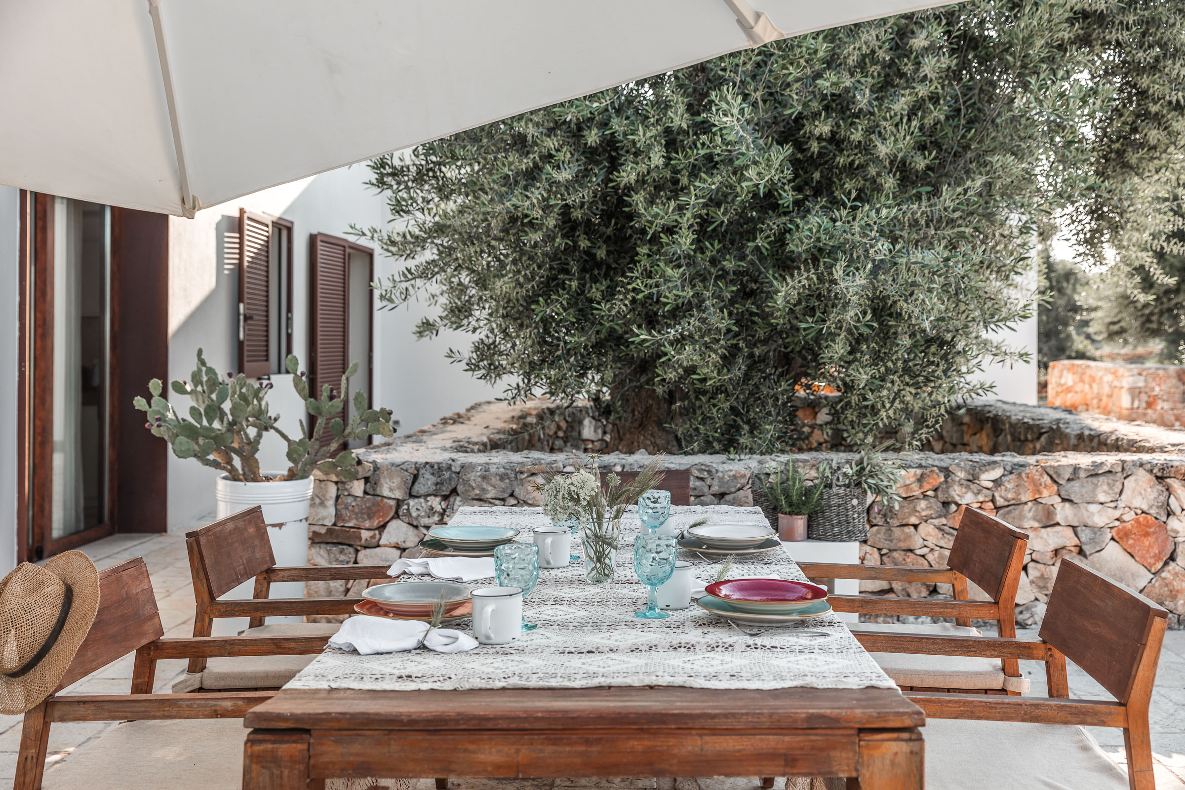 Mediterranean terrace dining area with wooden furniture, white plates and pink accents, climbing ivy and olive tree canopy overhead, stone walls